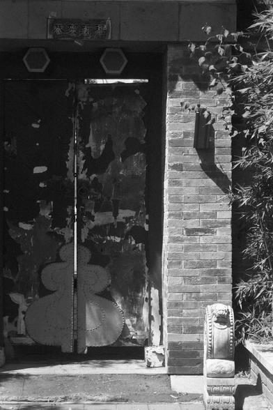 Ilford FP4 Plus 125 (FF)

English
A black-and-white photograph of a weathered wooden door with peeling paint, set in a brick wall. Above the door, there is a horizontal signboard with Chinese characters. The door features decorative metal elements, including two hexagonal shapes near the top and circular designs. A stone pillar with carved details stands to the right of the door, and foliage is visible on the right side of the frame.
中文
一张黑白照片，展示了一扇嵌入砖墙中的老旧木门，门上的油漆已脱落。门上方有一块水平的牌匾，上面写着几个中文字。木门上装…