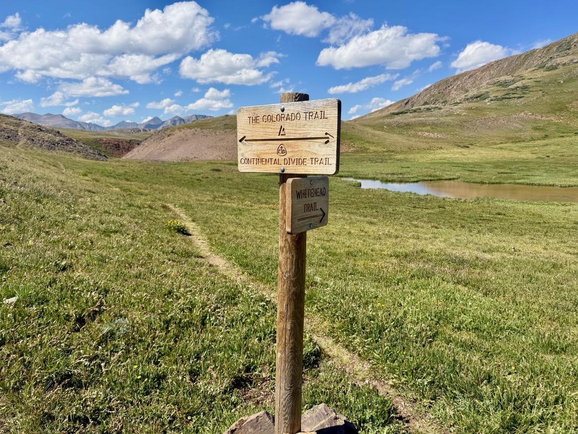 A tall trail sign stands in a green alpine tundra. It reads "The Colorado Trail" and "Continental Divide Trail" with arrows pointing both left and right. A smaller sign reads "Whitehead Trail" and points straight ahead. A faint trail leads ahead past a small pond with mountains on the horizon. Blue sky with puffy clouds.