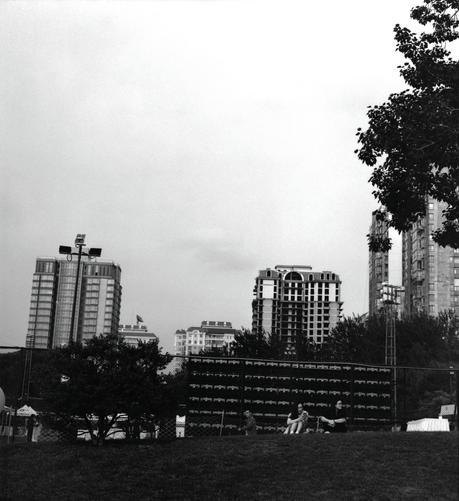Lucky LUCKY SHD 400 (6x7)

English
A black-and-white photograph of a park or recreational area. In the foreground, there is a grassy field with a few people sitting and relaxing. In the background, tall residential buildings rise against a cloudy sky. Trees and a fence are visible between the field and the buildings.
中文
一张黑白照片，展示了一个公园或休闲区。前景是一片草地，几个人坐着休息。背景是高层住宅楼，耸立在多云的天空下。草地和楼房之间可见树木和围栏。