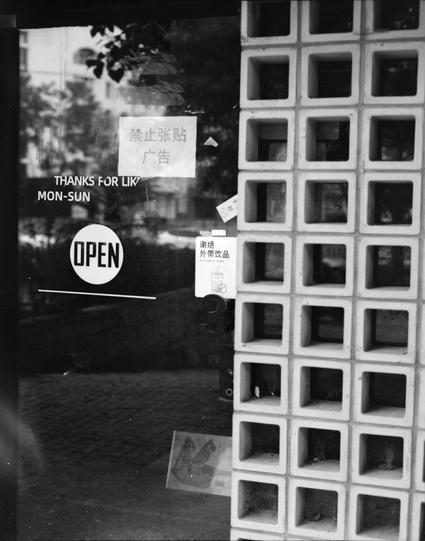 Lucky LUCKY SHD 400 (6x7)

English
A black-and-white photograph of a storefront window. The window displays several signs in both English and Chinese. The English text reads "THANKS FOR LIKE MON-SUN" and "OPEN" in a circular sign. The Chinese text includes "禁止张贴广告" (No Posting Advertisements) and "谢绝外带饮品" (No Outside Beverages). The window is adjacent to a wall made of square concrete blocks.
中文
一张黑白照片，展示了一家商店的橱窗。橱窗上贴有多个中英文标志。英文文字写着“THANKS FOR LIKE MON-SUN”和一个圆形“OPEN”标志。中文文字包括“禁止张贴广告”和“谢绝外带饮品”。…