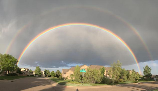 Photo showing full arc of a double rainbow above a sunlit street corner. A street sign shows the names of intersecting streets, Mourning Dove and Prairie Ridge. Gray clouds overhead are dripping virga, precipitation that doesn’t reach the ground because the lower air is so dry. Colorado, June 2017.