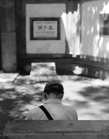 Lucky LUCKY SHD 100 (6x7)

English
A black-and-white photograph of a person sitting on a wooden bench, facing away from the camera. The person wears a cap and a shoulder bag. In front of them is a signboard with Chinese characters and additional smaller text below. The scene is set in a park-like environment with trees casting dappled shadows on the ground.
中文
一张黑白照片，展示一个背对镜头坐在木椅上的人。该人戴着帽子，背着一个肩包。他们面前有一个标志牌，上面写着模糊的中文字以及下方的小字。场景设定在一个公园般的环境中，树木在地面上投射出斑驳的阴影。