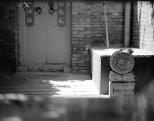 Lucky LUCKY SHD 100 (6x7)

English
A black-and-white photograph of a traditional courtyard. The scene features a closed wooden door with two vertical decorative panels on either side, each adorned with circular designs. The door is set within a brick wall, with a tiled floor leading up to it. To the right, there is a stone pedestal and a large, cylindrical stone carving with intricate patterns. Sunlight casts sharp shadows, illuminating parts of the courtyard and creating a contrast between lig…