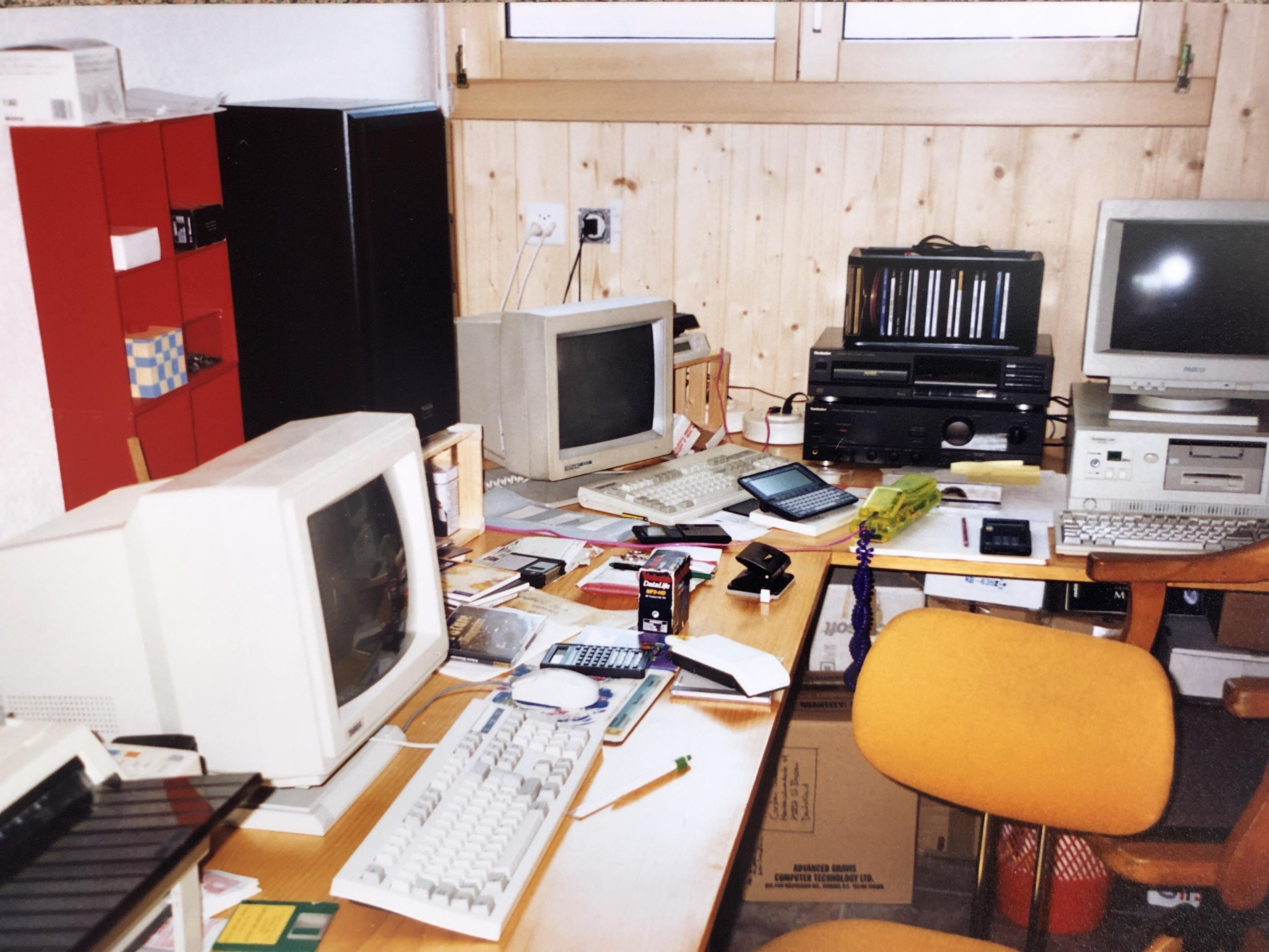 Photo of corner desk with 3 CRT monitors. The desks are littered with paper, pens, calculators, phones, diskettes, ..
