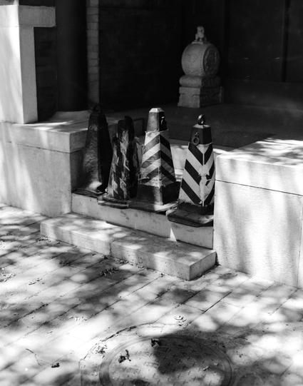 Lucky LUCKY SHD 100 (6x7)

English
A black-and-white photograph showing a set of steps leading up to a wooden door. On the doorsteps, there are several traffic cones, some of which are black and white striped. The steps are made of light-colored stone or concrete. In the background, there is a stone pillar with a decorative top. Shadows from nearby trees or objects are visible on the steps and platform.
中文
一张黑白照片，展示了一组木门前的台阶。平台上放置着多个交通锥，其中一些是黑白相间的条纹。台阶由浅色石头或混凝土制成。背景中有一石头的装饰，顶部装饰精美。台阶和平台上可以看到附近树…