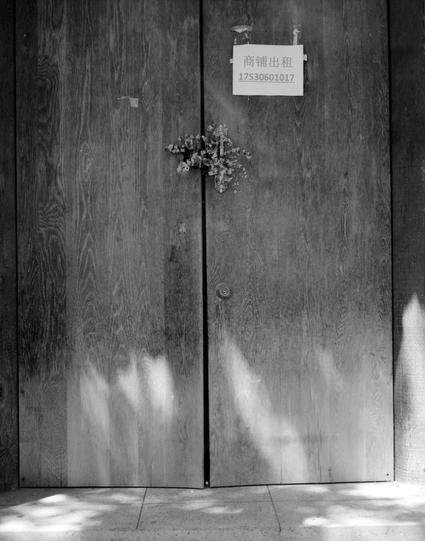 Lucky LUCKY SHD 100 (6x7)

English
A black-and-white photograph of a wooden double door with a visible wood grain pattern. A small piece of dried plant is attached to the left door. Above the doors, there is a rectangular sign with Chinese text that reads "商铺出租" (Shop for Rent) followed by a phone number: 17530601017. Shadows from nearby trees or objects are visible on the doors.
中文
一张黑白照片，展示了一扇木质双开门，门上有明显的木纹图案。左侧门上有干的植物。门上方挂有一个矩形标志，上面用中文写着“商铺出租”，后面跟着一个电话号码：17530601017。门上可以看到附近树木或物体的影子。