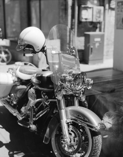 Lucky LUCKY SHD 100 (6x7)

English alt text: Black-and-white close-up photo of a motorcycle in display. The front of the motorcycle is turned slightly to the left. A round headlight and a clear windshield or windscreen stand in front of the handlebars and instrument area. The motorcycle has a round side mirror on the left side. The front wheel and tire tread are visible at the bottom, with the wheel angled slightly toward the camera. In the background, shelves or containers and other display it…