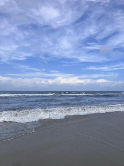 A serene beach scene featuring gentle waves lapping against the shore under a partly cloudy blue sky.