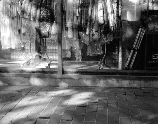 Lucky LUCKY SHD 100 (6x7)

English
A black-and-white photograph of a storefront window display. The window features numerous garments, mostly dresses, covered in plastic wraps and hanging on racks. Some clothing items are also placed on a metal rack inside the store. The pavement in front of the store is made of rectangular bricks, and sunlight casts shadows of the window frames onto the ground.
中文
一张黑白照片，展示了一家商店的橱窗陈列。橱窗内悬挂着许多用塑料膜包裹的服装，大多是连衣裙，挂在衣架上。店内还有一个金属衣架，上面也放着一些衣物。商店前的人行道由长方形砖块铺成，阳光将窗框的影子投…