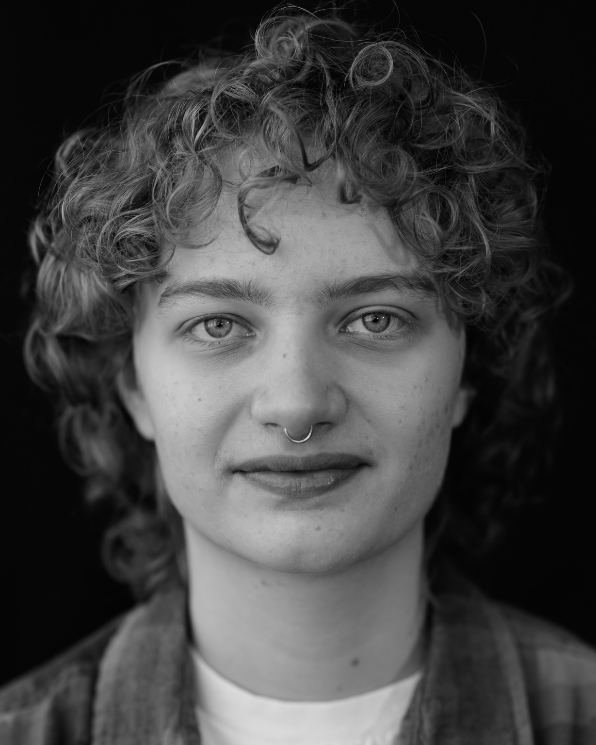 A black and white headshot of a young adult: they are looking directly at the lens with bright eyes, curly, tousled hair, and a simple nose ring.
