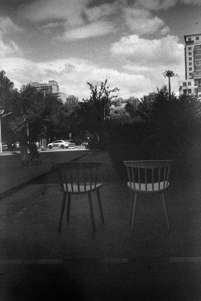 ERA 100 (FF)

English
A black-and-white photograph showing two empty chairs in the foreground, placed on a paved surface. In the background, there is a street with trees lining both sides, buildings, and a few vehicles. The sky is partly cloudy, and the scene appears to be taken from a slightly elevated perspective.
中文
一张黑白照片，前景是两把空椅子，放置在铺砌的地面上。背景是一条两侧种有树木的街道，还有建筑物和几辆车。天空部分多云，场景看起来是从一个稍高的角度拍摄的。