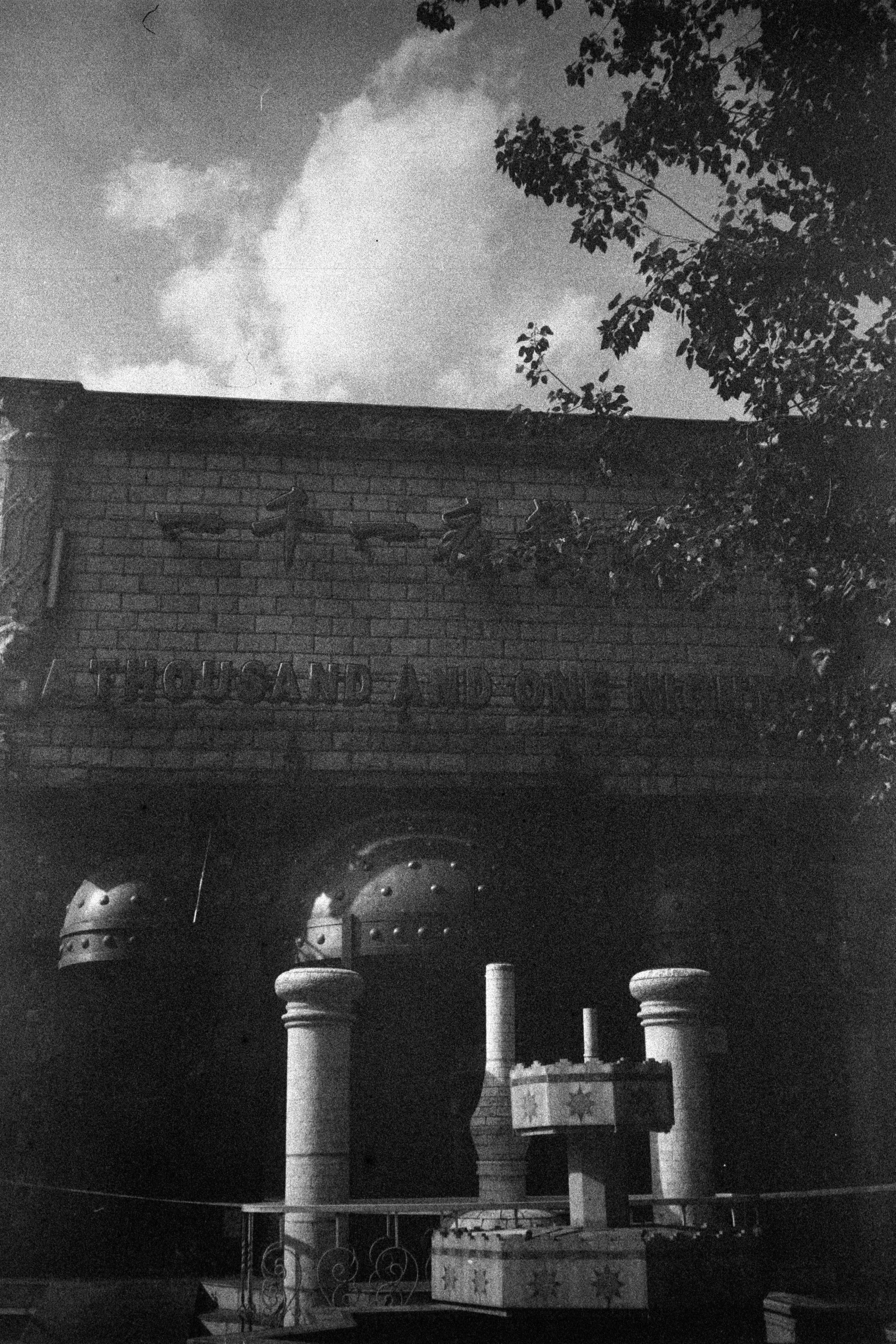 ERA 100 (FF)

English
A black-and-white photograph of a brick structure with the text "THOUSAND AND ONE NIGHTS" carved at the top. Below the text, there are decorative elements resembling a crescent moon and stars. The structure features ornate pillars and a circular design with geometric patterns. Trees are visible on the right side of the image, and the sky is partially cloudy.
中文
一张黑白照片，拍摄的是一座砖结构建筑，顶部雕刻着“ONE THOUSAND AND ONE NIGHTS”文字。文字下方有新月和星星的装饰图案。建筑物前方有装饰性的柱子和一个带有几何图案的圆形设计。图片右侧可见树木，天空部分多云。