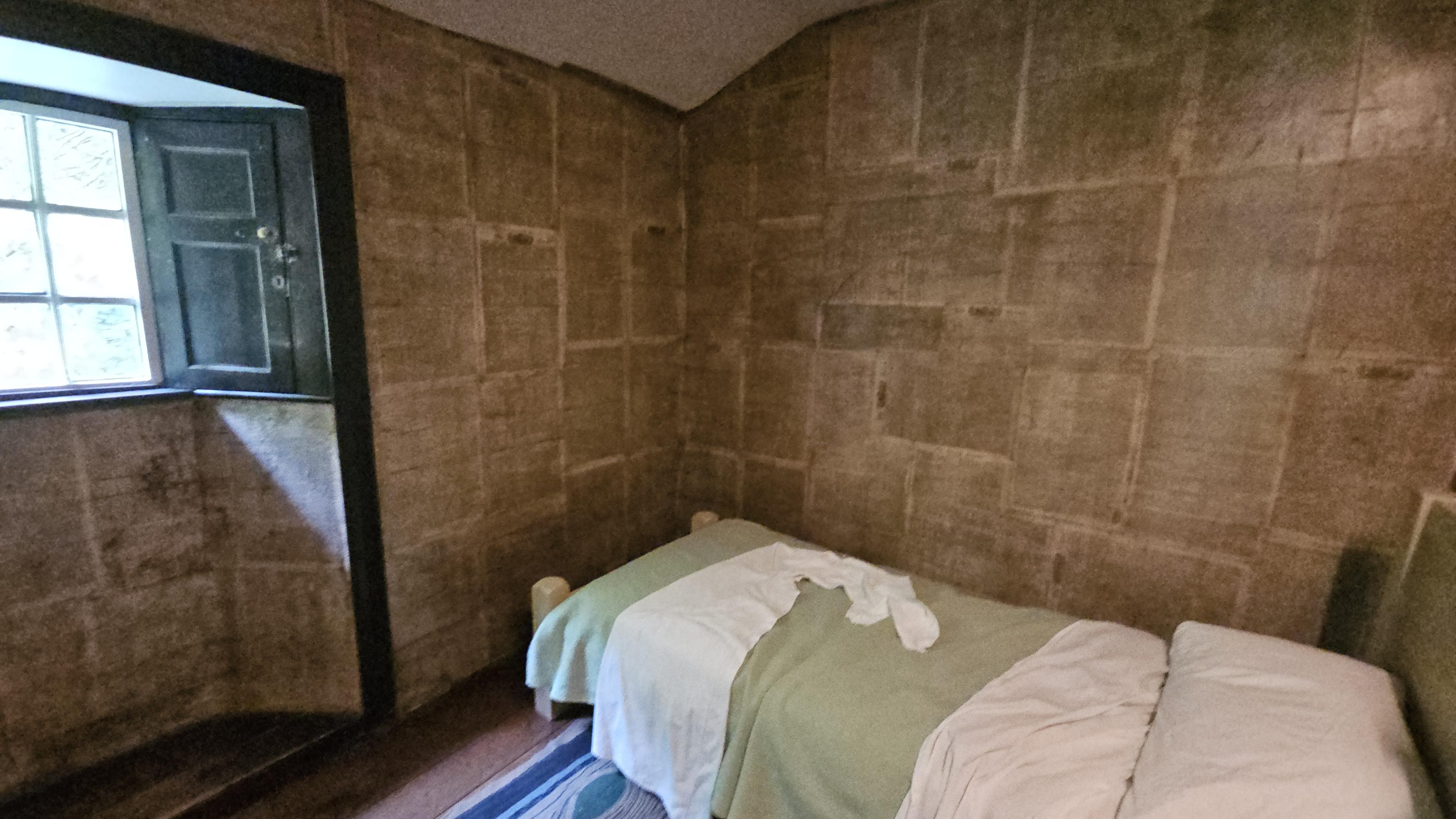 Interior of a small bedroom. There is a single bed and a window, and the walls are covered with 19th Century newspapers.