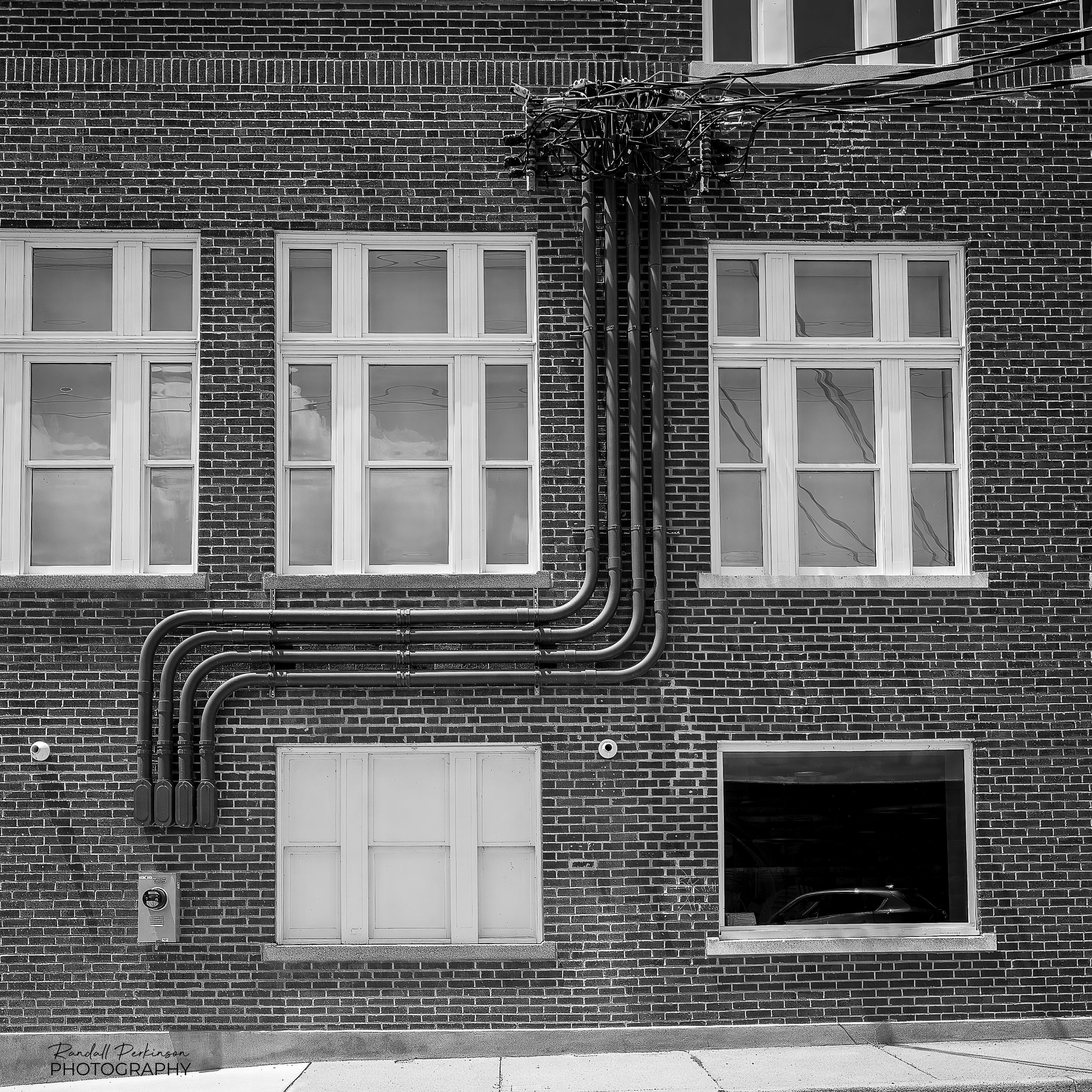 A brick building with multiple windows has four large electrical conduits that drop down from where the wires come to the building, turn 90 degrees to left and travel horizontally between two windows, and then turn 90 degrees down before entering the building.