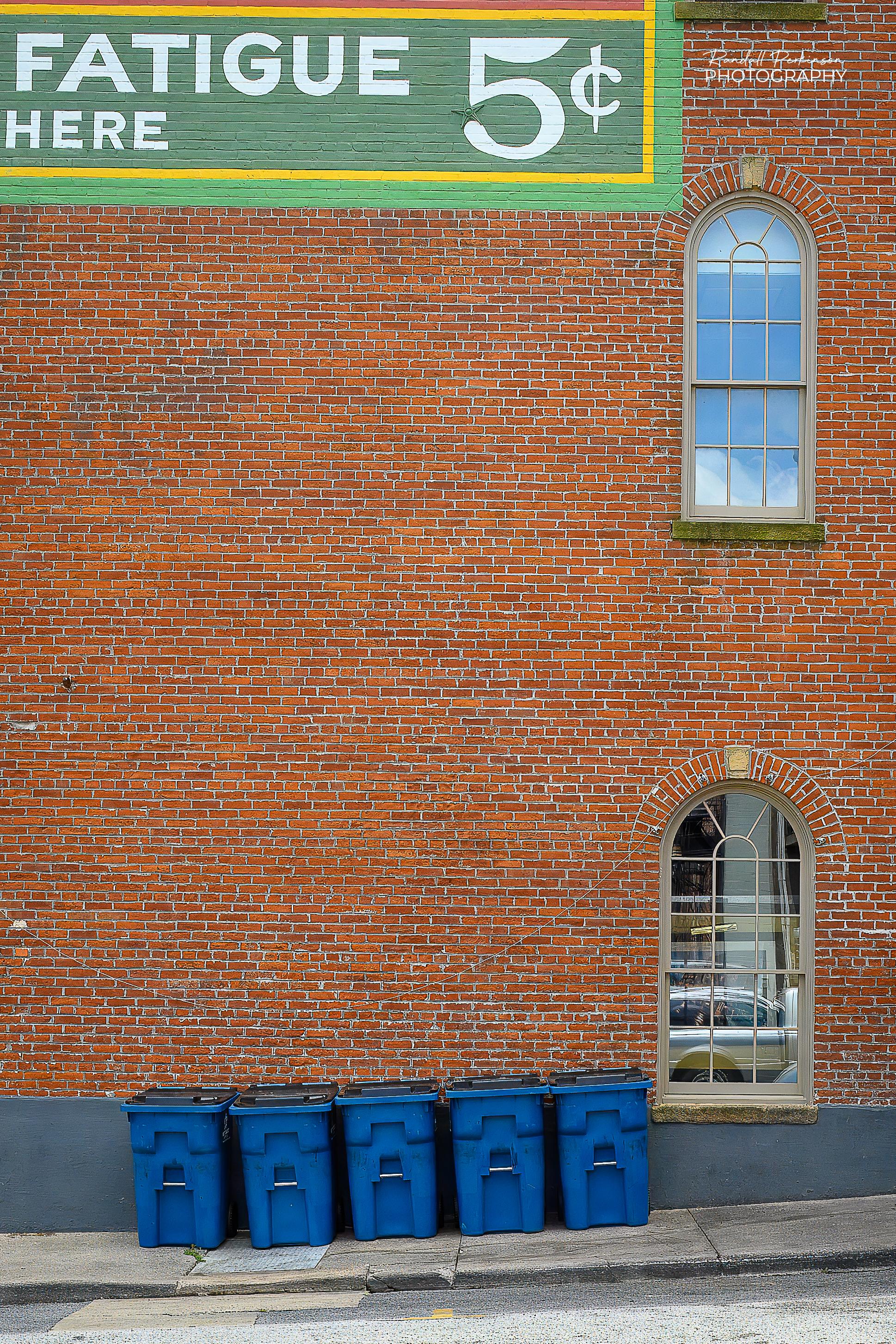 Five blue trash carts with black lids sit side by side on a sidewalk by a multi-story brick building.  Part of a painted sign appears in the upper left showing the words "Fatigue" and "here" and "5 cents".