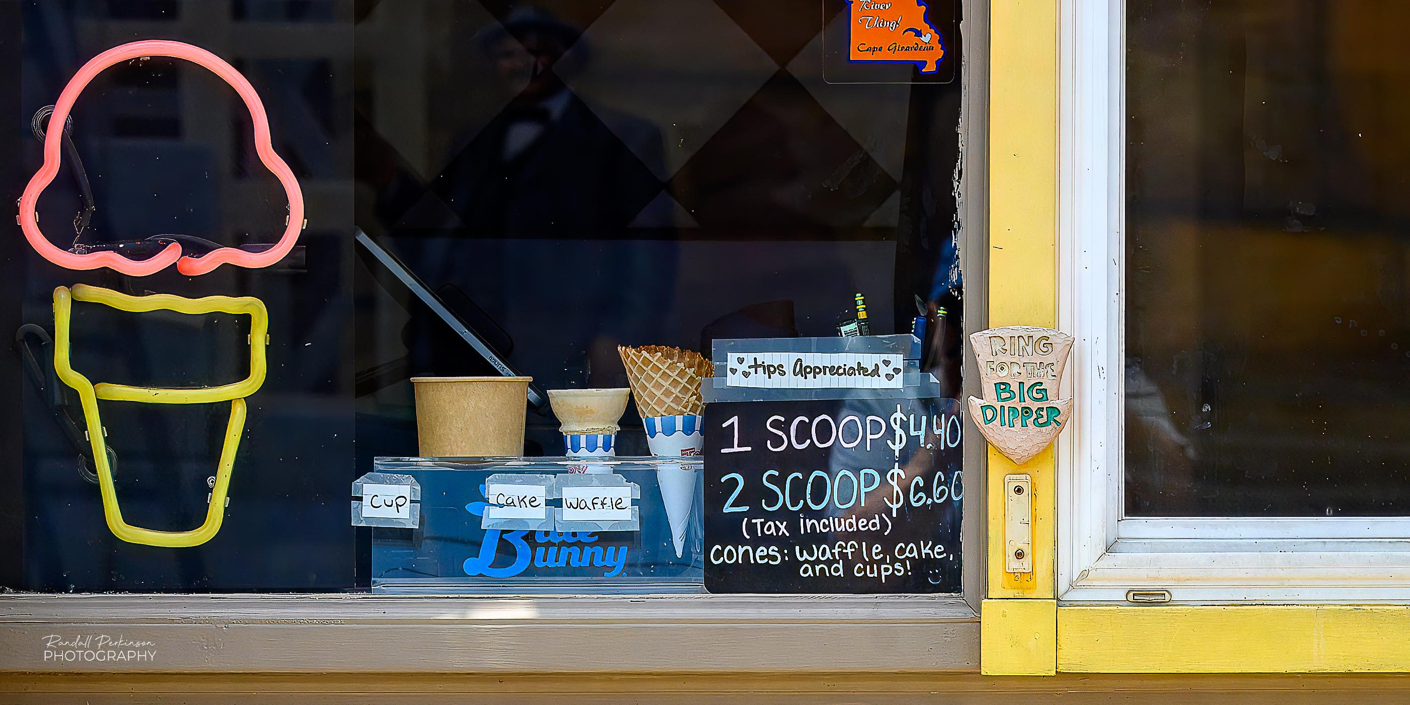 A view of a ice cream store street window with a neon sign of an ice cream cone, a display of the different cup and cone options, a sign showing the ice cream prices, and a doorbell with a sign that says "Ring for the Big Dipper".
