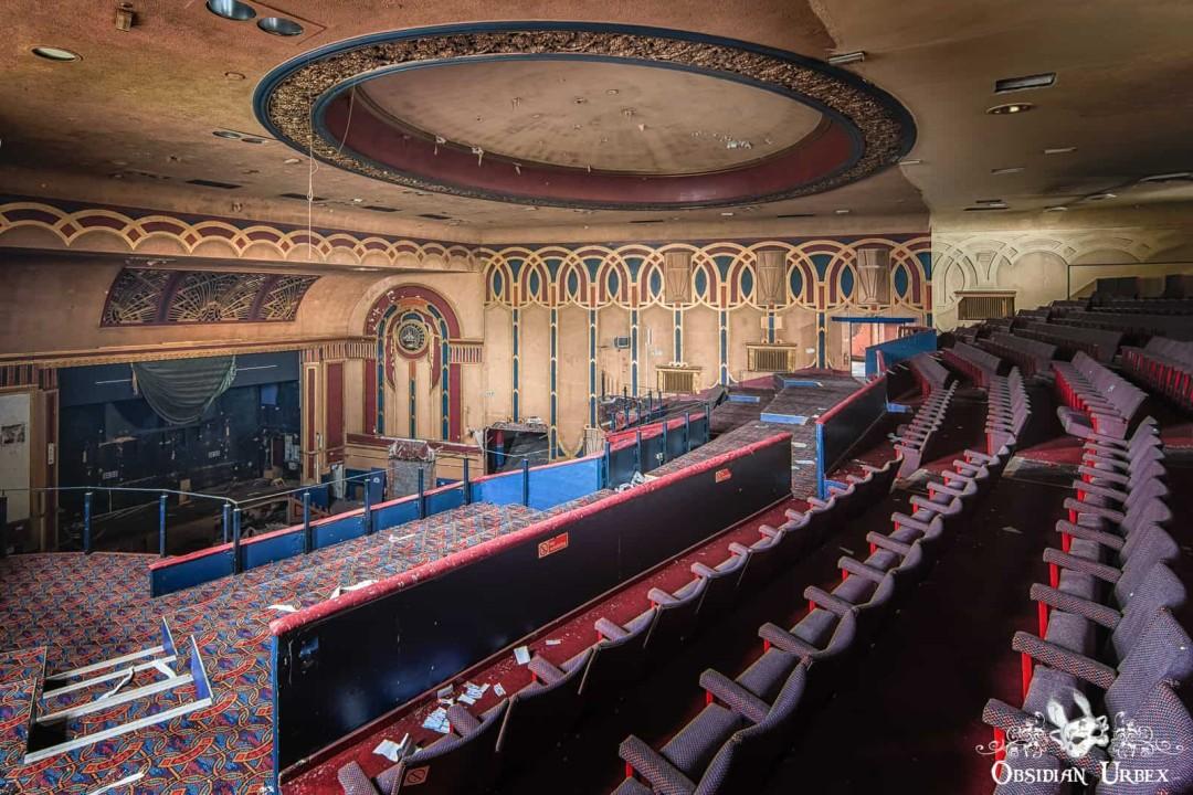 A grand, decaying auditorium features rows of red seating and an ornate ceiling above a stage area. Detailed wall patterns add to its faded glory