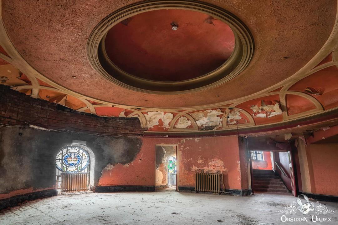 An abandoned room, likely in an old cinema, features a round domed ceiling, peeling red paint, and a stained-glass window.