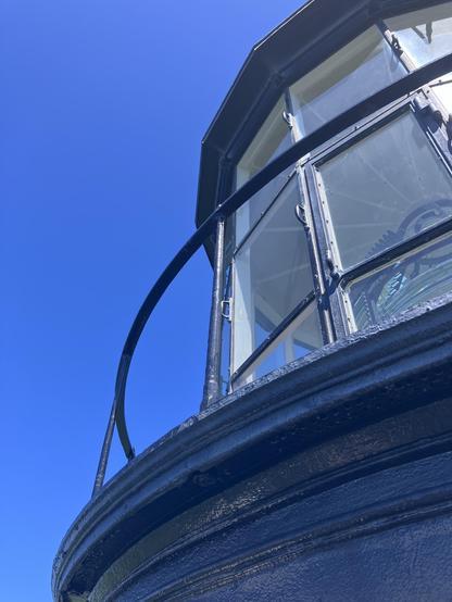 Close-up view of a lighthouse lantern house against a clear blue sky. The structure features glass panels and a black metal railing.