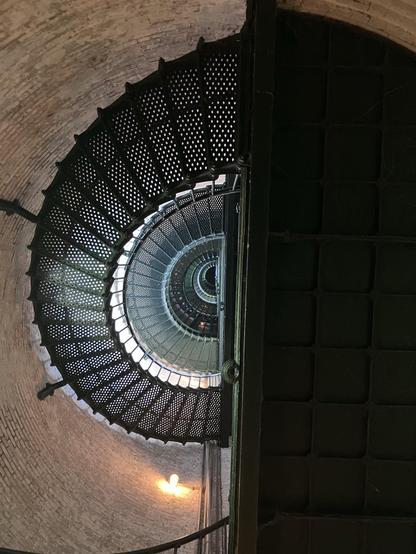 Looking up from the base of a spiral metal staircase towards the distant top of the lighthouse. The outer circular walls are made of brick. 