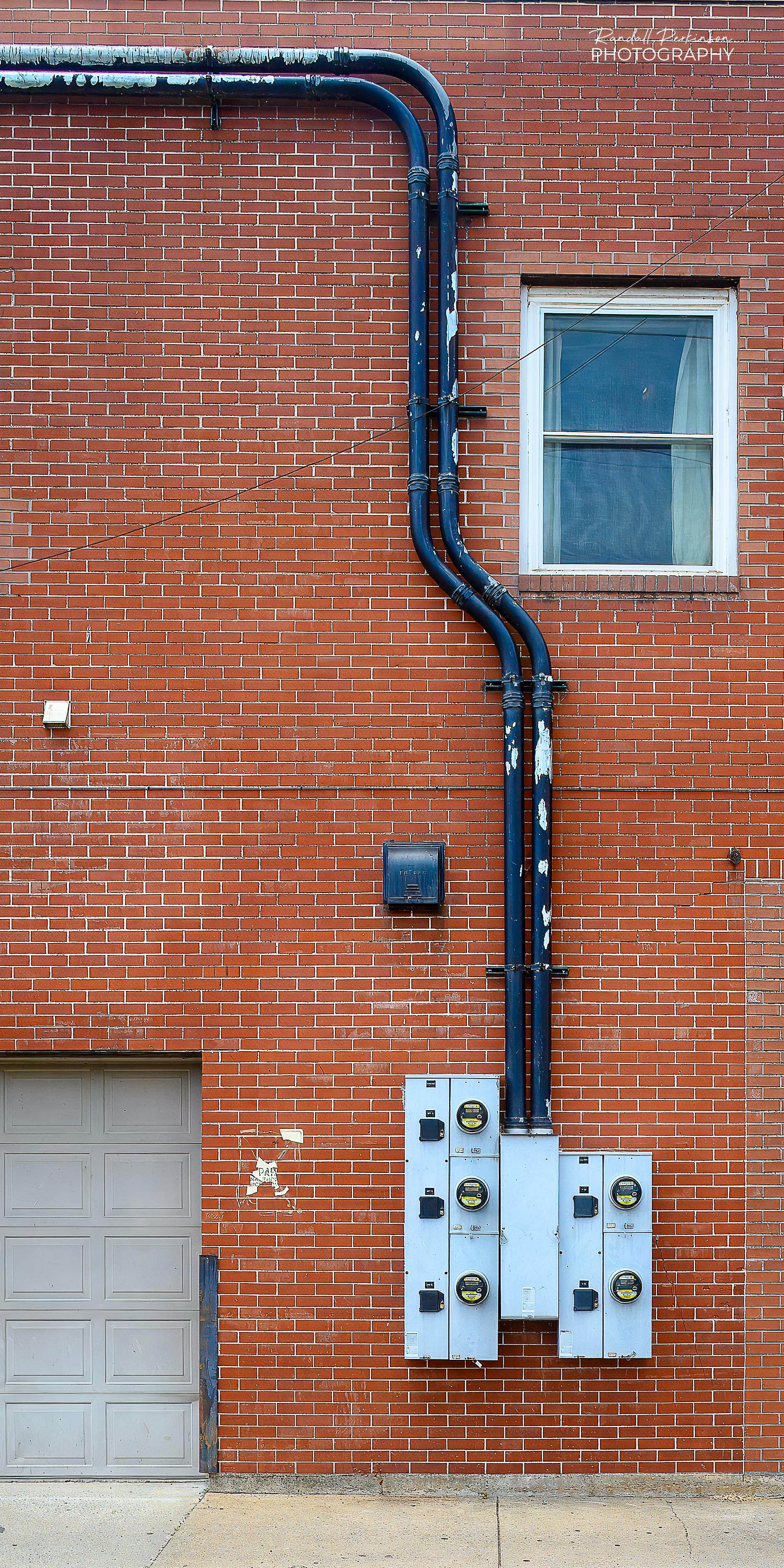Two large electrical conduits run across and down the face of a multi-story brick building to 5 electric meters serving tenants in the building.