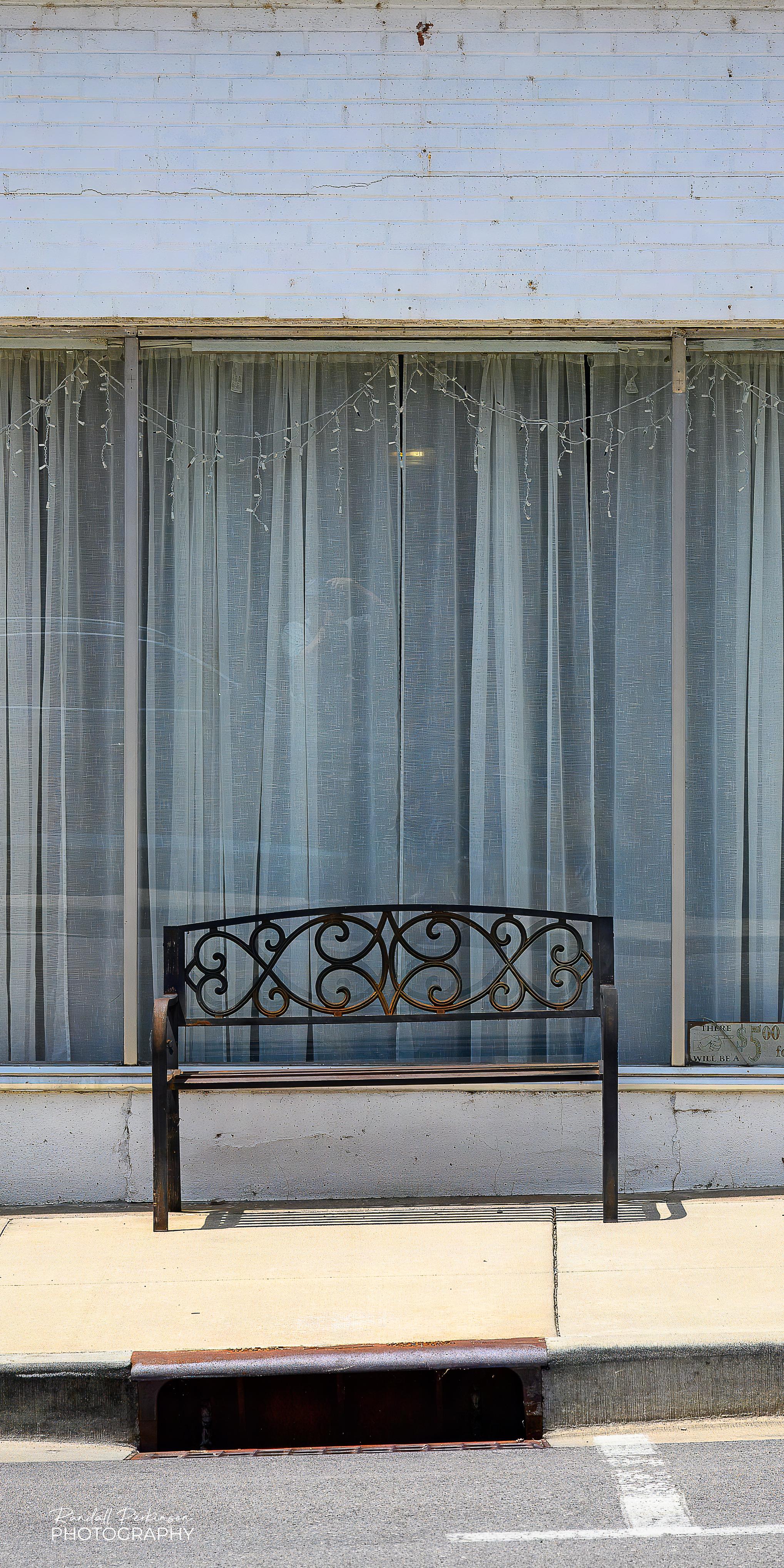 A single black bench with a decorative back sits on the sidewalk in front of a large window with curtains waiting for somebody to come by and use it.
