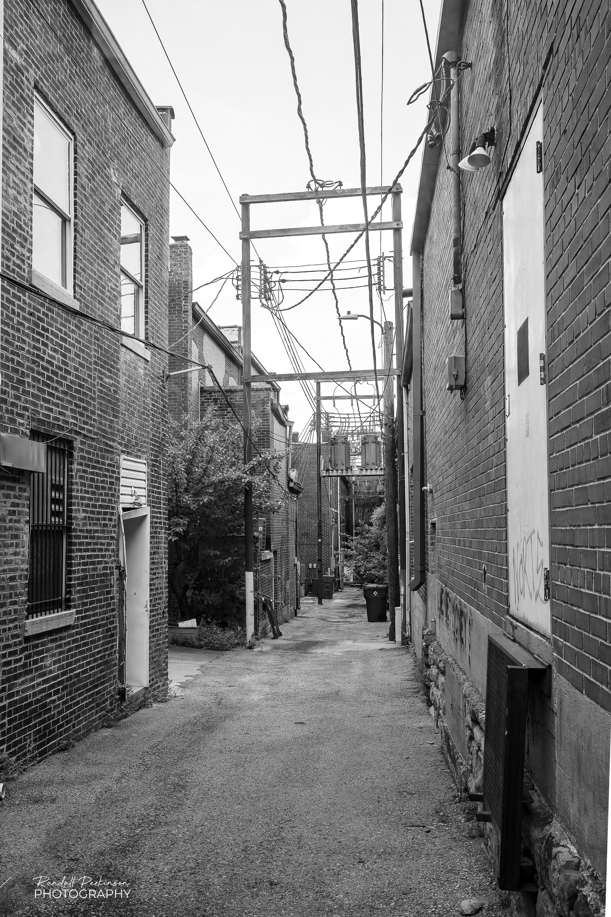 A narrow empty alley provides access to a number of old two story buildings.  Electrical lines are strung on multi-leg wooden frames that span the alley.