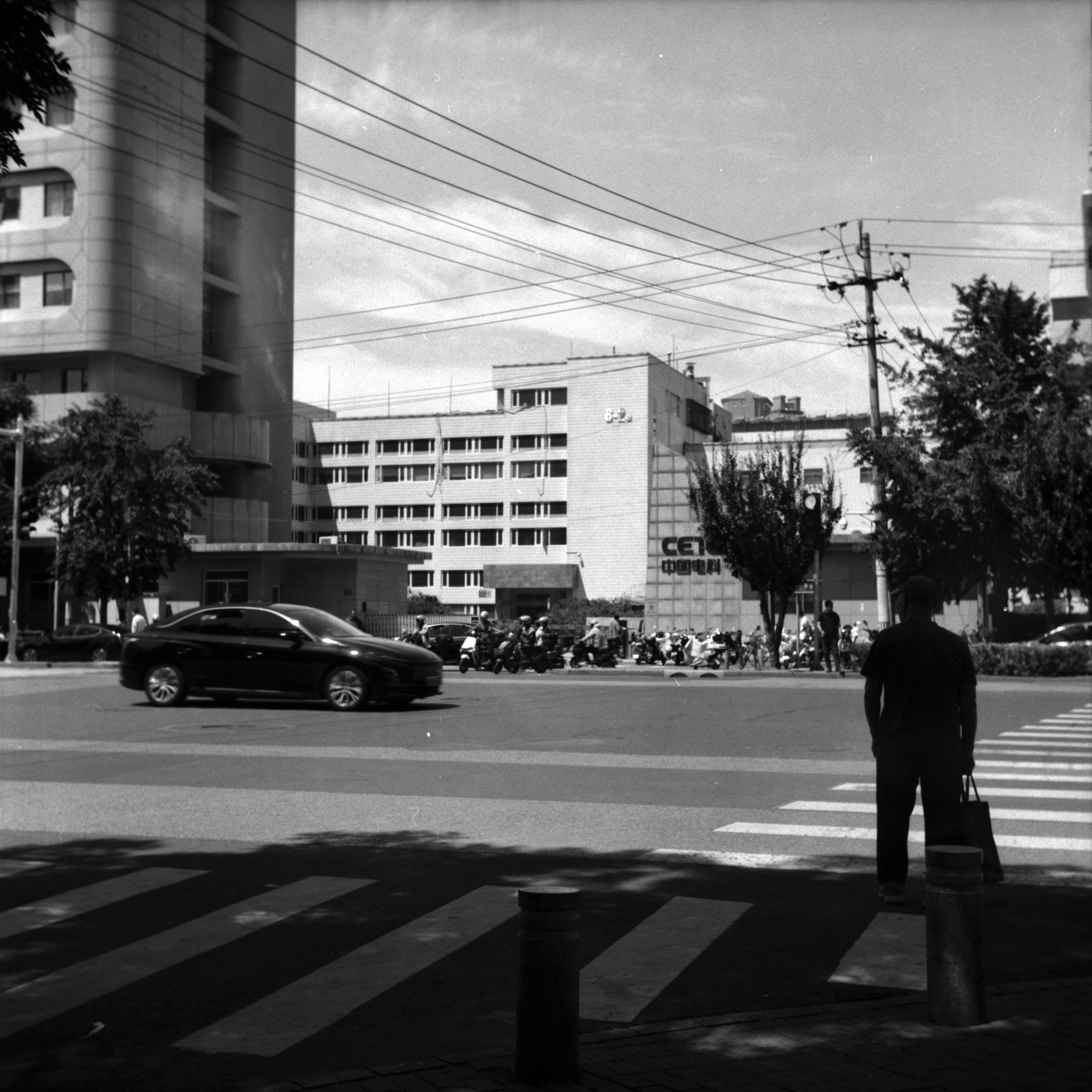 Lucky LUCKY SHD 100 (6x6)

English
A black-and-white photograph of an urban street scene. A person stands on the right side of the frame, facing away from the camera, near a pedestrian crossing. The street is busy with a dark-colored car driving in the middle of the road. In the background, there are multiple buildings, including a tall, curved building on the left and a mid-rise building. Trees line the street, and power lines crisscross above. Several motorbikes and people are visible near the buildings.
中文
一张黑白城市街道的照片。右侧有一个人背对着镜头站在人行横道附近。街道上有一辆深色汽车行驶。背景中有多栋建筑，左侧是一栋高大的弧形建筑，中间有一栋中等高度的建筑。街道两侧种有树木，上方有交叉的电线。建筑附近还可以看到多辆摩托车和行人。
