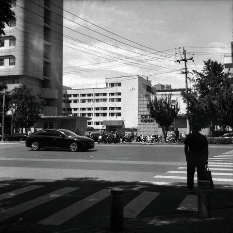 Lucky LUCKY SHD 100 (6x6)

English
A black-and-white photograph of an urban street scene. A person stands on the right side of the frame, facing away from the camera, near a pedestrian crossing. The street is busy with a dark-colored car driving in the middle of the road. In the background, there are multiple buildings, including a tall, curved building on the left and a mid-rise building. Trees line the street, and power lines crisscross above. Several motorbikes and people are visible near th…