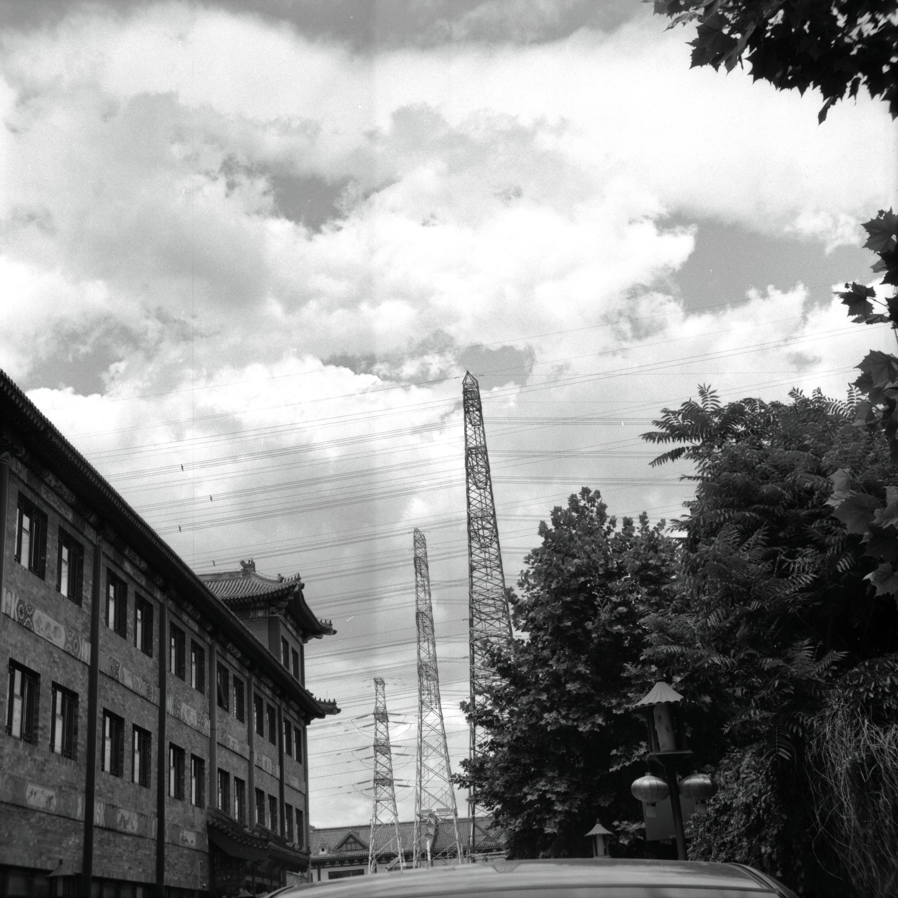Lucky LUCKY SHD 100 (6x6)

English
A black-and-white photograph featuring a traditional multi-story brick building on the left side of the frame. The building has ornate details and a tiled roof. In the background, there are tall metal lattice towers, likely electrical pylons, rising into a cloudy sky. Trees and foliage partially obscure the view of the towers and the building.
中文
一张黑白照片，左侧是一栋传统的多层砖结构建筑，建筑上有装饰性细节和瓦片屋顶。背景中有高大的金属格子塔，可能是电力铁塔，耸入多云的天空。树木和植被部分遮挡了塔和建筑的视线。