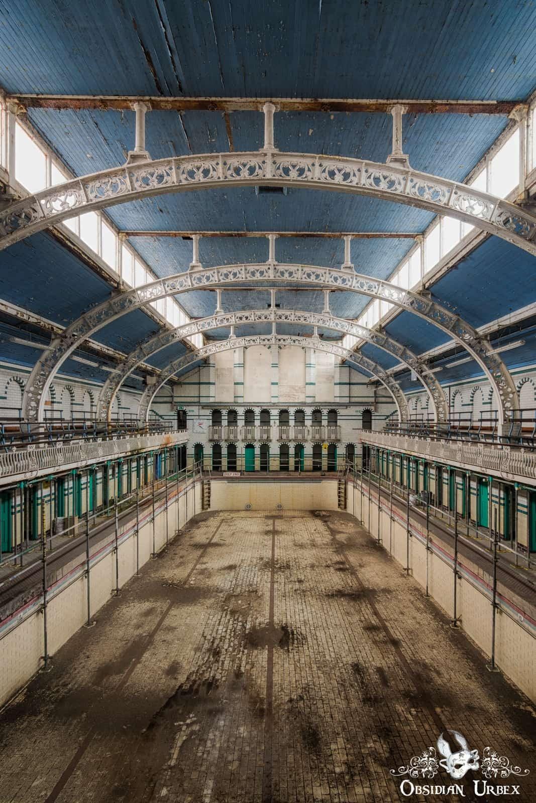 An abandoned indoor swimming pool, with a vast empty basin, blue ceiling, and ornate white arched supports.