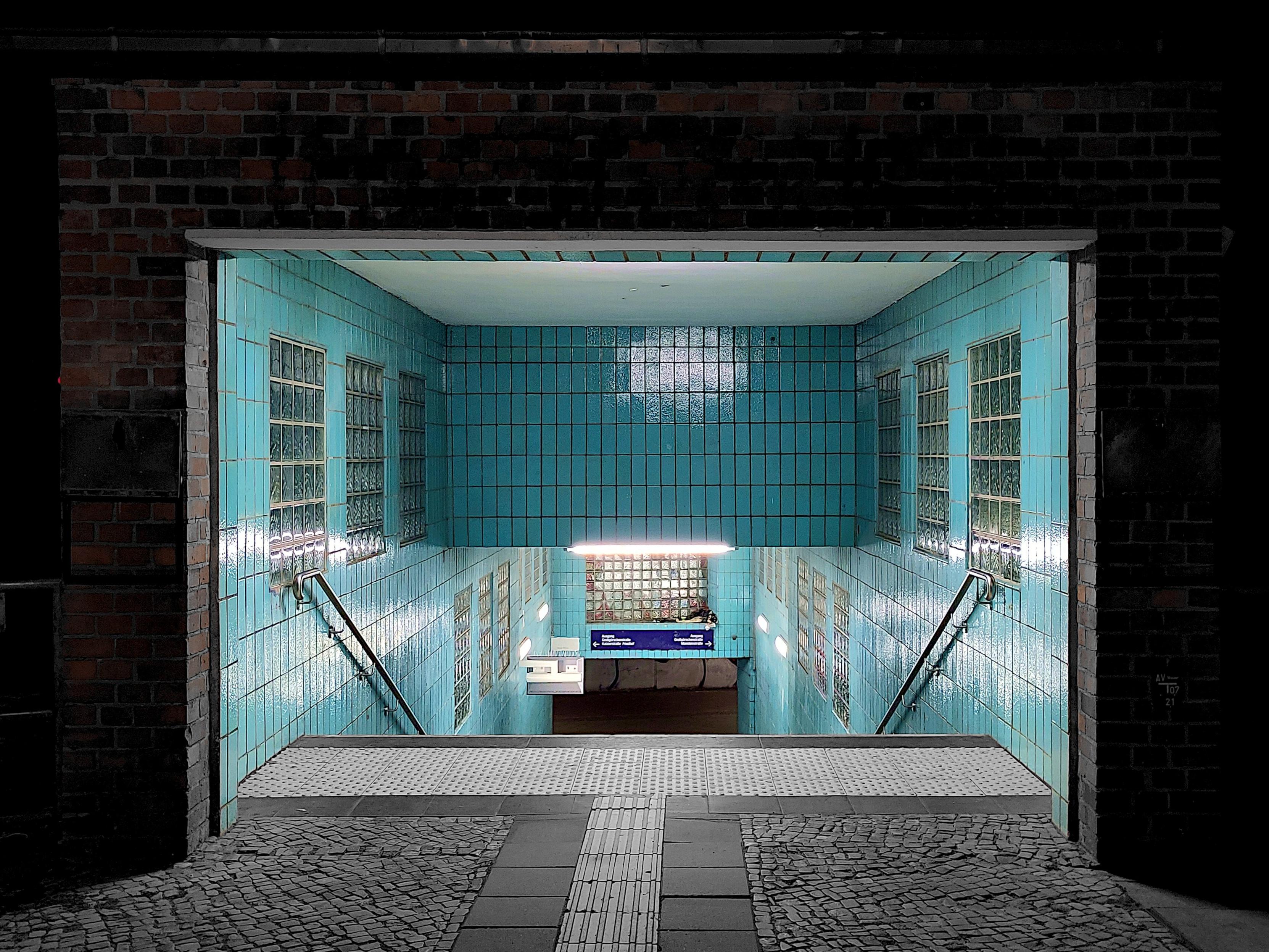 A nighttime view of a subway station entrance set into a dark brick wall. A staircase descends underground between walls lined with turquoise tiles and glass-block windows, brightly lit by fluorescent tubes. Metal handrails run down both sides. At the bottom, a blue directional sign with white text and arrows is mounted on the tiled wall. The foreground is a paved surface with cobblestones and a strip of tactile paving leading to the top of the stairs. The tiled interior glows blue-green against the surrounding darkness.