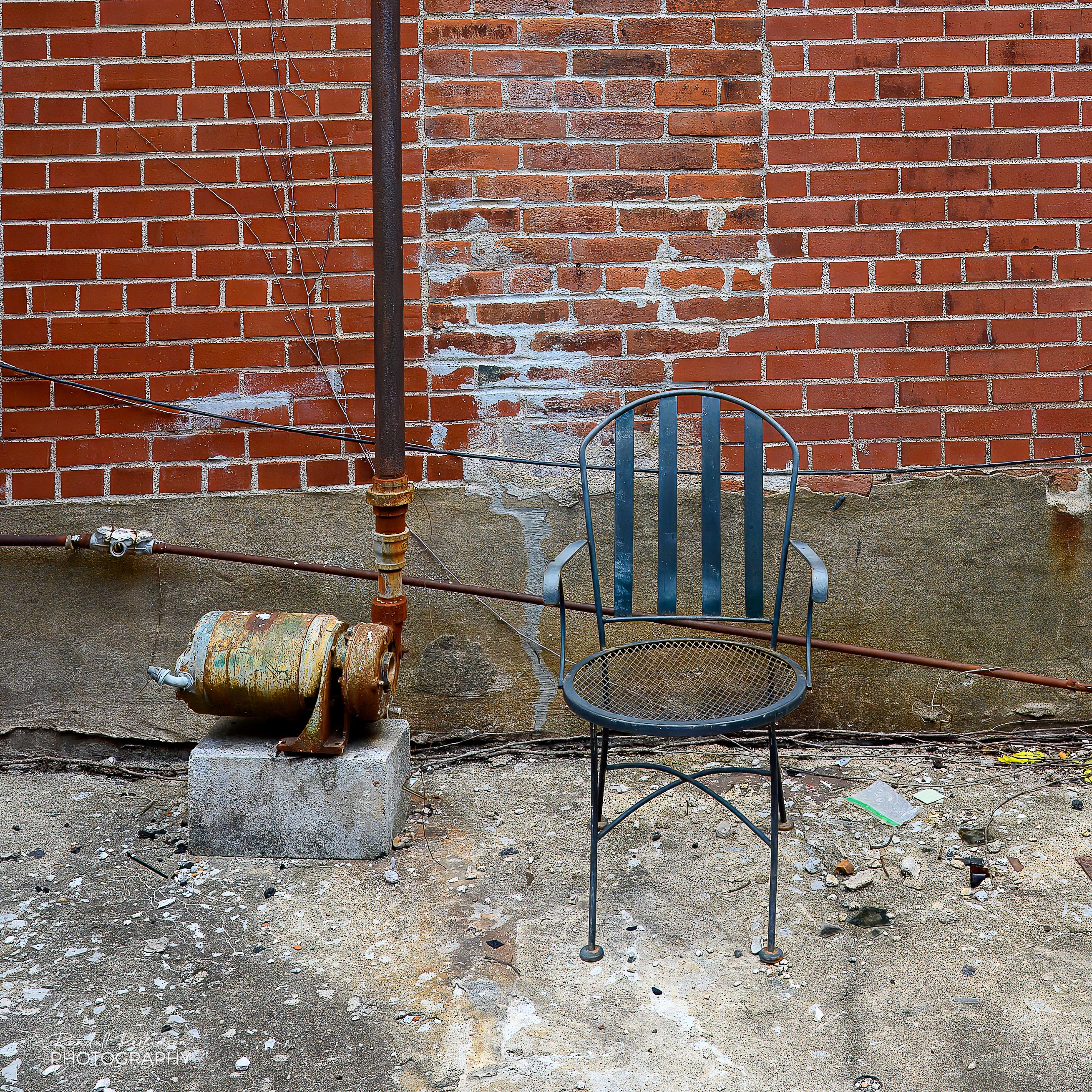A metal chair sits in an alley in front of a brick wall.  Next to it is a rusty old pump with a discharge pump going vertical out of it.  The pump has had it's electrical connection removed from a drooping conduit running along the lower part of the wall.