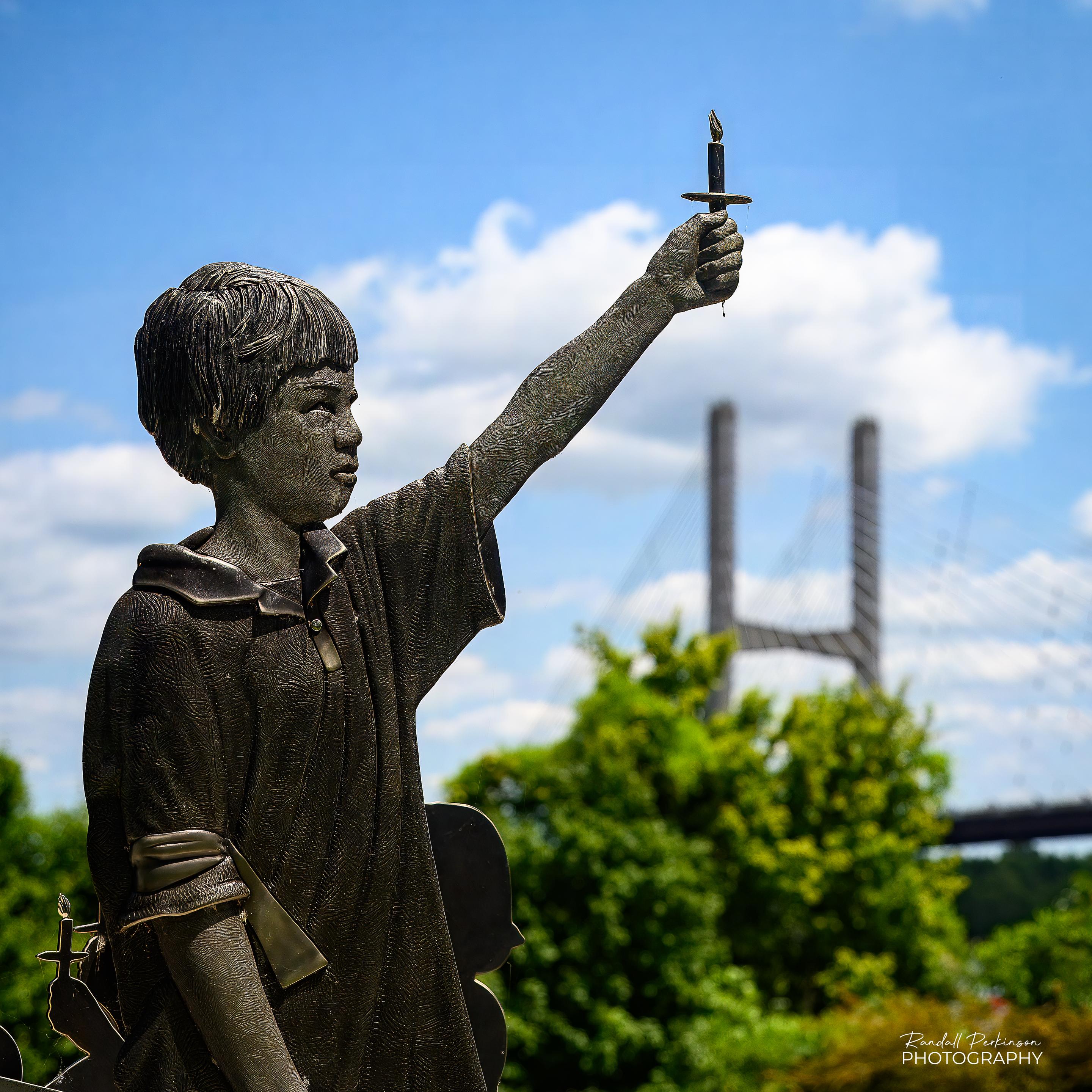 This boy holding a candle raised in the air is part of a larger sculpture by an unknown artist in Riverfront Bridge Park in Cape Girardeau, Missouri.  In the background you can see trees and one of the tall bents of the cable stayed bridge over the Mississippi River.