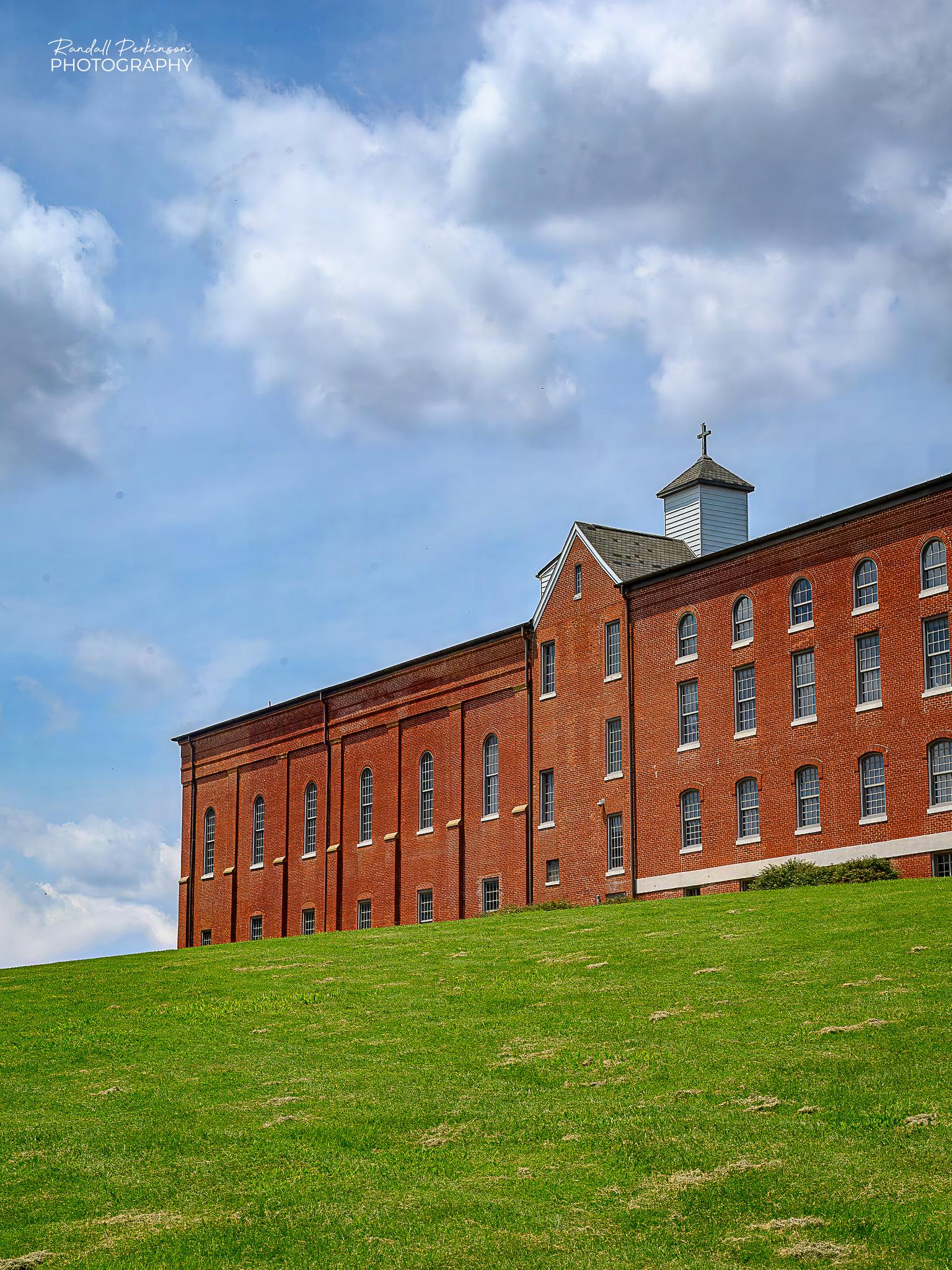 A large multi-story bridge building sits at the top of a grass hill under blue skys and clouds.  The building is part of the Southeast Missouri State University's River Camps that houses all of the fine arts programs.  The campus orignated as St. Vincent's College which was closed in 1979 and later bought by SEMO.