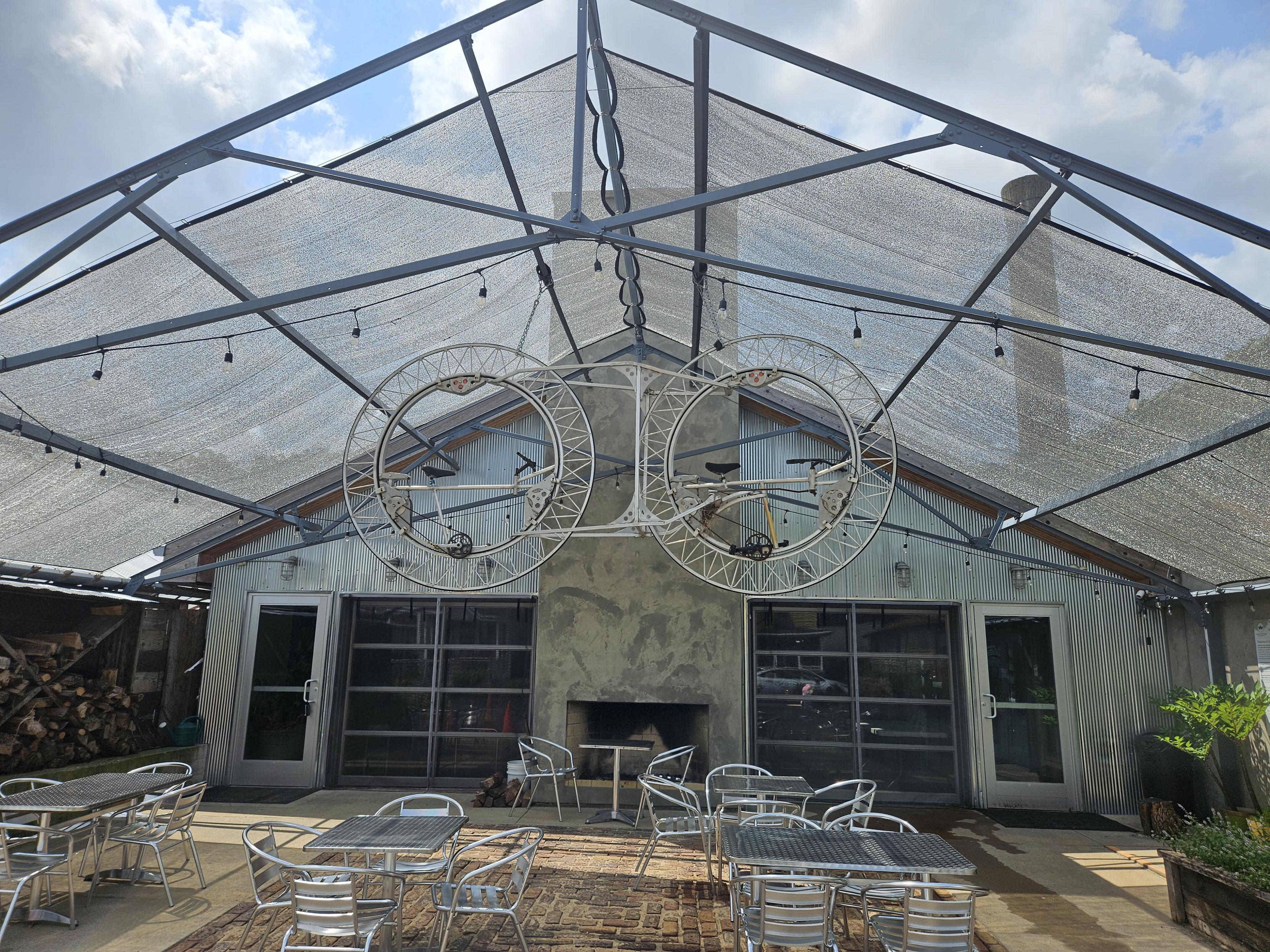 A covered patio with tables and chairs. Above thise is a sculpture that resemble bicycle wheels with bicycle Saddles and chains on the inside of the wheels