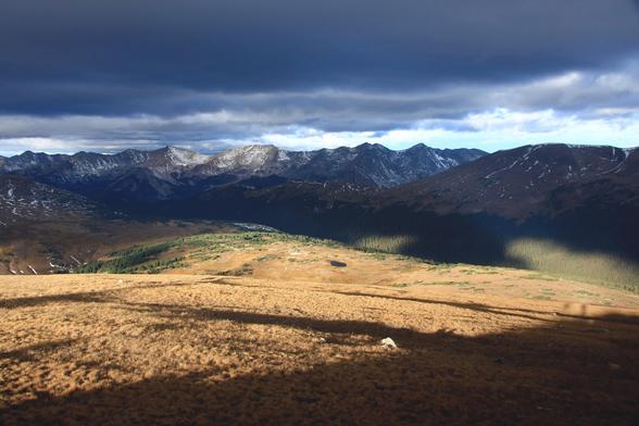 Photo of the Never Summer Range in the Rocky Mountains, looking to the west from Trail Ridge road in Rocky Mountain National Park. Just past sunrise on a September morning, most of the range is still in the shadow of mountains behind and around me, except for a small section just left of the center of the image. The foreground is brightly sunlit tundra with a few shadows of people standing near me stretching across the sparse brown vegetation. Grey clouds span the sky above a narrow streak of b…