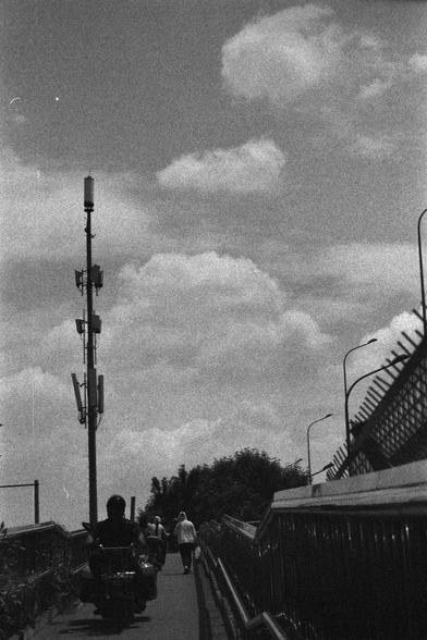 Ilford HP5 Plus 400 (FF)

English
A black-and-white photograph of a bridge or walkway. A tall utility pole with multiple antennas and equipment is prominently positioned on the left side of the frame. The sky is partly cloudy, with large, soft clouds scattered across it. A few people are walking along the path, and a railing runs parallel to the walkway on the right side.
中文
一张黑白照片，拍摄的是一座桥或步道。左侧有一根高大的公用事业杆，上面安装了多个天线和设备。天空部分多云，散布着大块柔和的云朵。几个人在小路上行走，右侧有一个栏杆与步道平行。