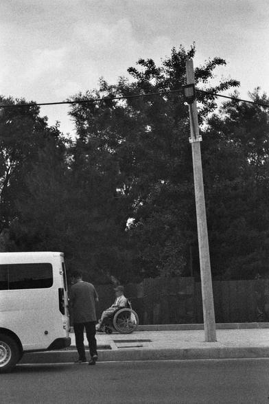 Ilford HP5 Plus 400 (FF)

English
A black-and-white photograph showing a street scene. A white van is parked on the left side of the frame. A person stands near the van, facing away from the camera. In the background, another individual is seated in a wheelchair on the sidewalk, near a tall utility pole. Dense trees form the backdrop, and the sky is overcast.
中文
一张黑白照片，展示了一个街道场景。左侧停着一辆白色厢式车。一个人的背影站在厢式车附近。背景中，另一个人坐在轮椅上，位于人行道上，靠近一个高大的公用事业杆。背景是茂密的树木，天空阴云密布。