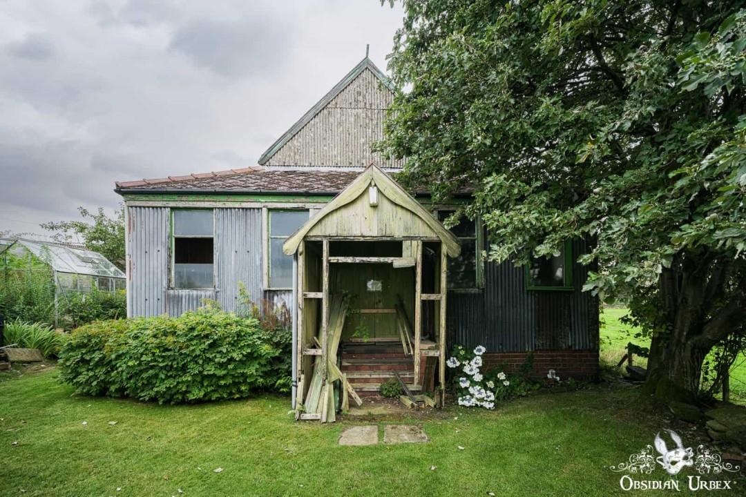 An old, weathered building with a small porch and corrugated siding stands in an overgrown garden under an overcast sky