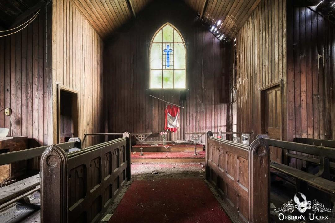 An abandoned wooden church interior with a red carpet aisle leading to a stained-glass window featuring a blue cross. Wooden pews line the sides