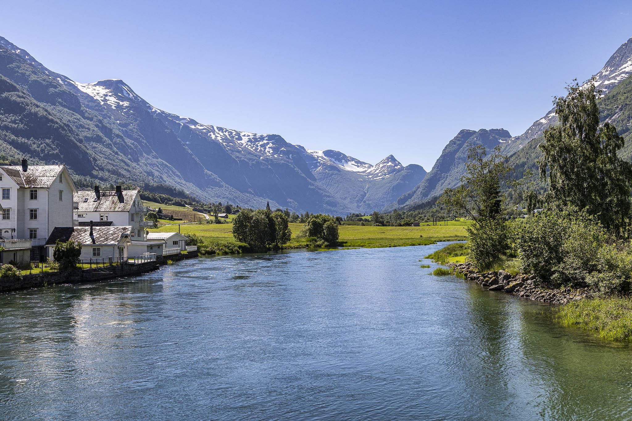 It's a beautiful, clear, warm, sunny day, and we're within the fjordland area of Norway in the middle of summer, visiting a small town. We're standing on a low road bridge over a river where it joins a fjord around which the houses and businesses of this picturesque location have sprung up, and we're looking inland to where the mountains rise in the distance to either side of this split in the landscape. The water is flat and motionless, reflecting much of the surrounding scenery. It's wide beneath us, stretching beyond the left and right sides of view, but heads away, narrowing and curving around to the right out of sight behind a grassy bank of land dotted with bushes and trees at the water's edge. On the left side of the river there are half a dozen buildings, most low but one of them over three storeys high, or four if you count the window in the attic space. All the buildings are covered in white wooden slats, glowing against the greens and blues around, and all have tiling on the