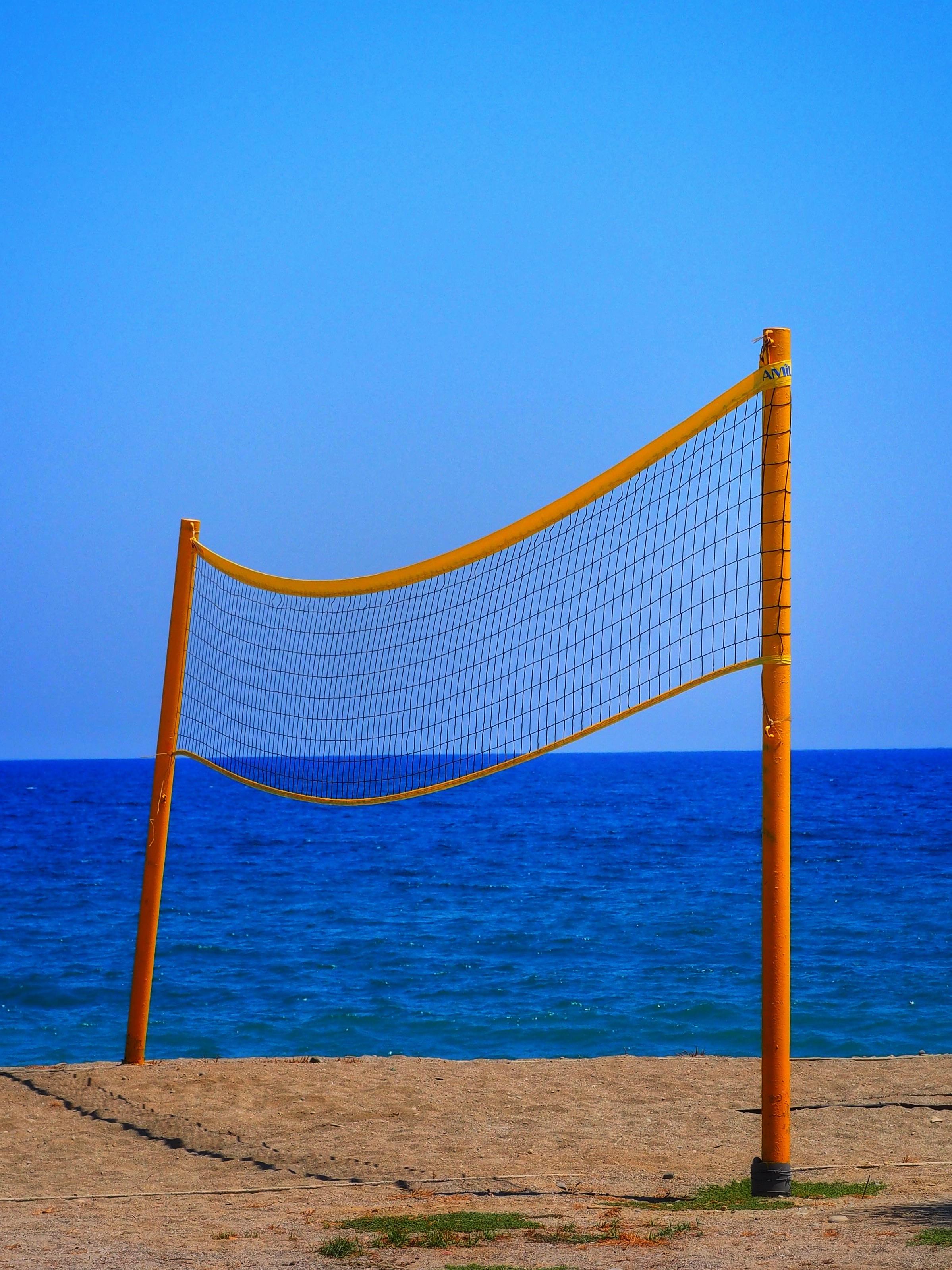 A yellow beach volleyball net stands on sandy ground, with a clear blue sky and the sea in the background.