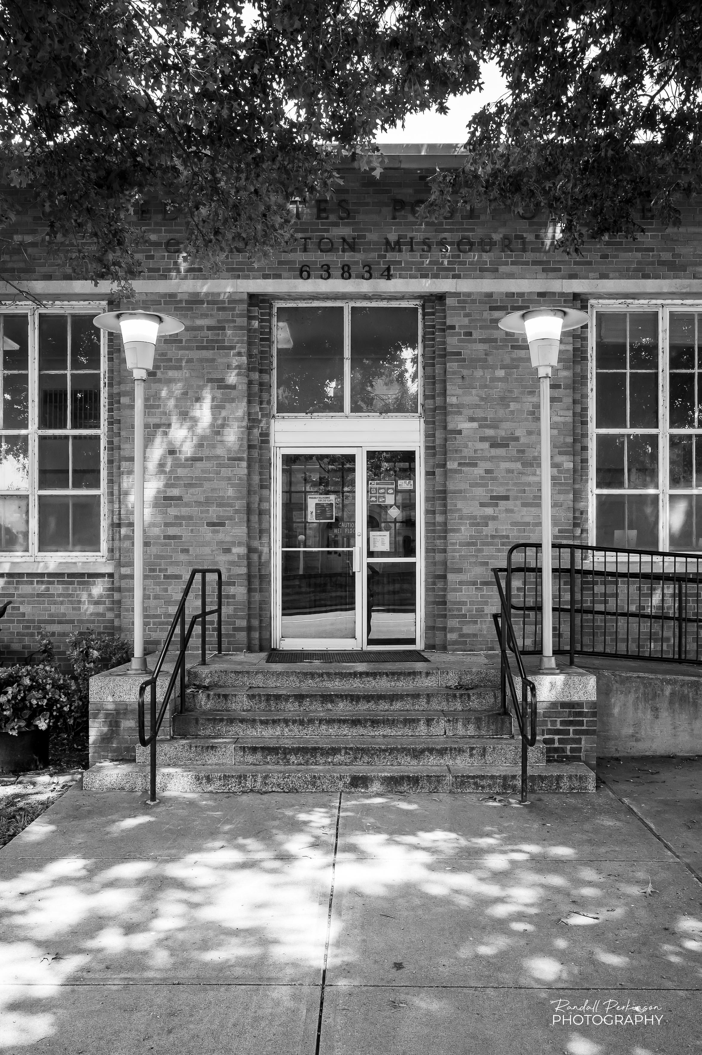 A view of the front entrance of the United States Post Office in Charleston, Missouri.  The brick building has tall windows flanking the entrance door and two light posts on either side of steps leading to the door.