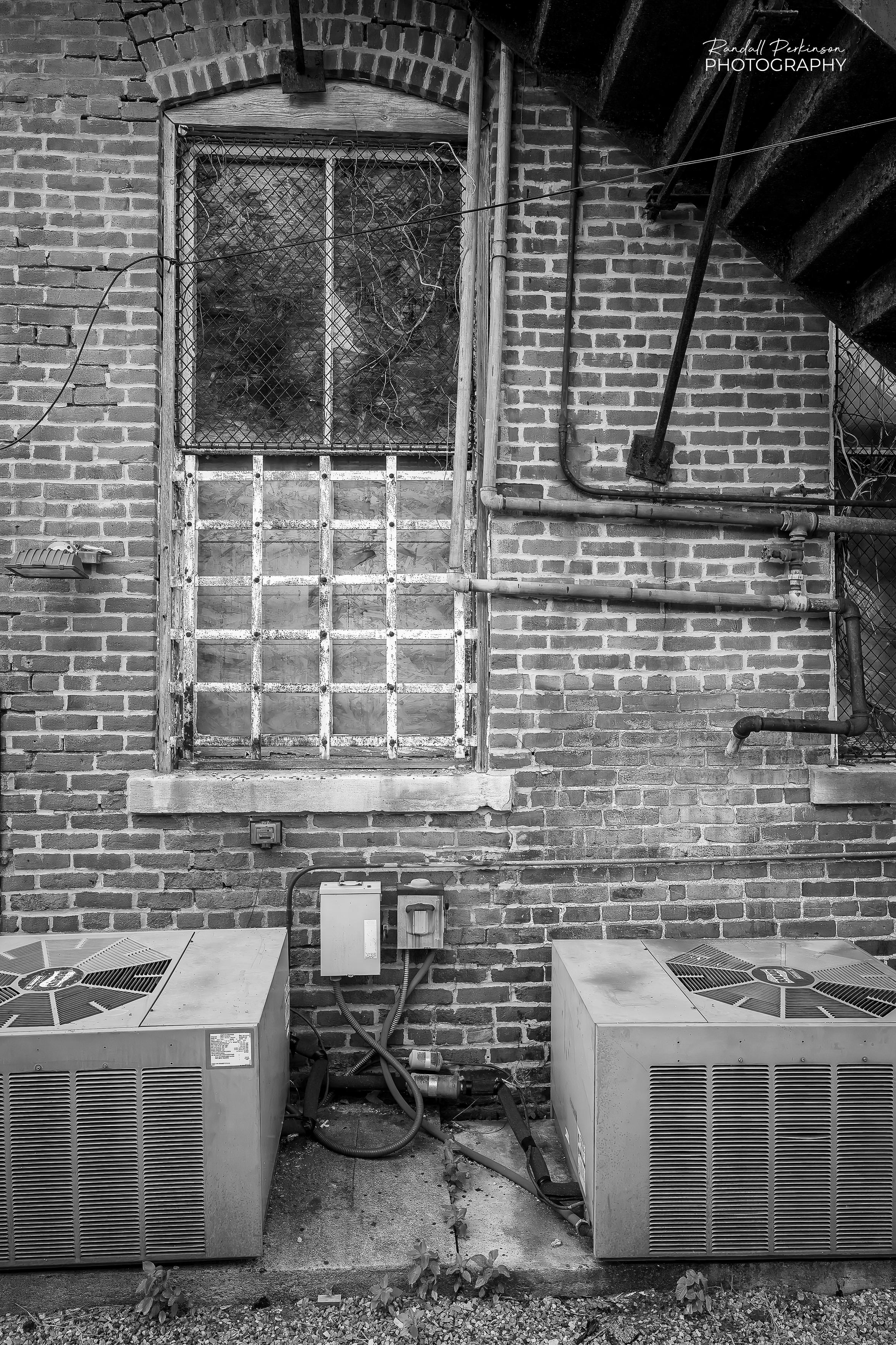 A black and white view of a window on the back of a brick building located under a metal staircase.  The window has bars and wire over it to keep people out.  There are two air conditioning units sitting on a concrete pad undere the window.