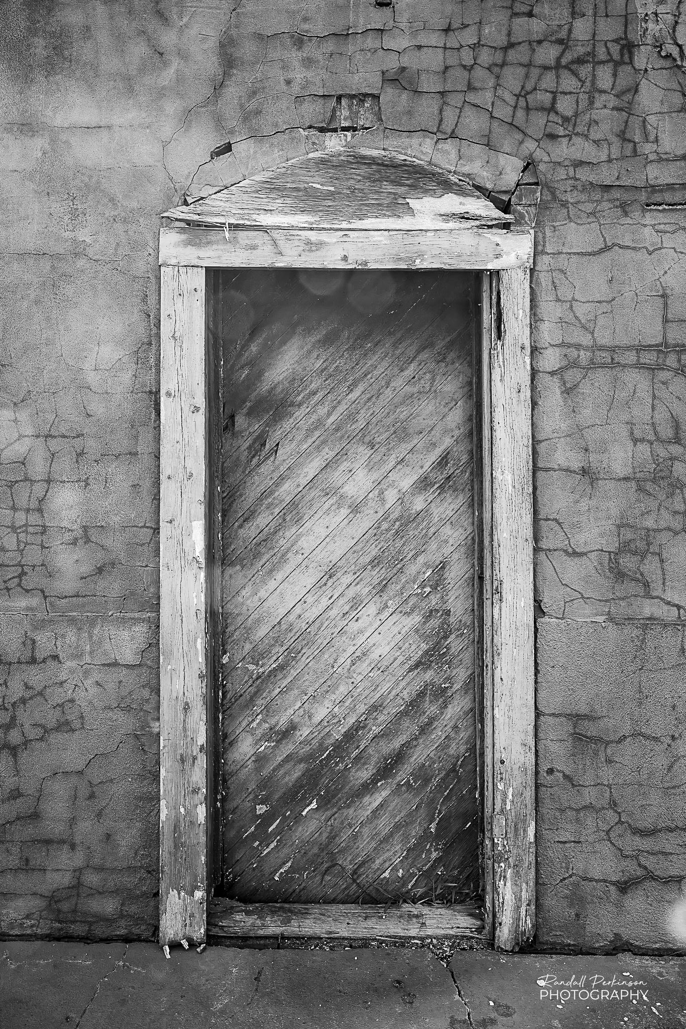 A wooden door made of diagonal boards sits inside a rough wood frame in a wall with plaster/cement coating is highly cracked.