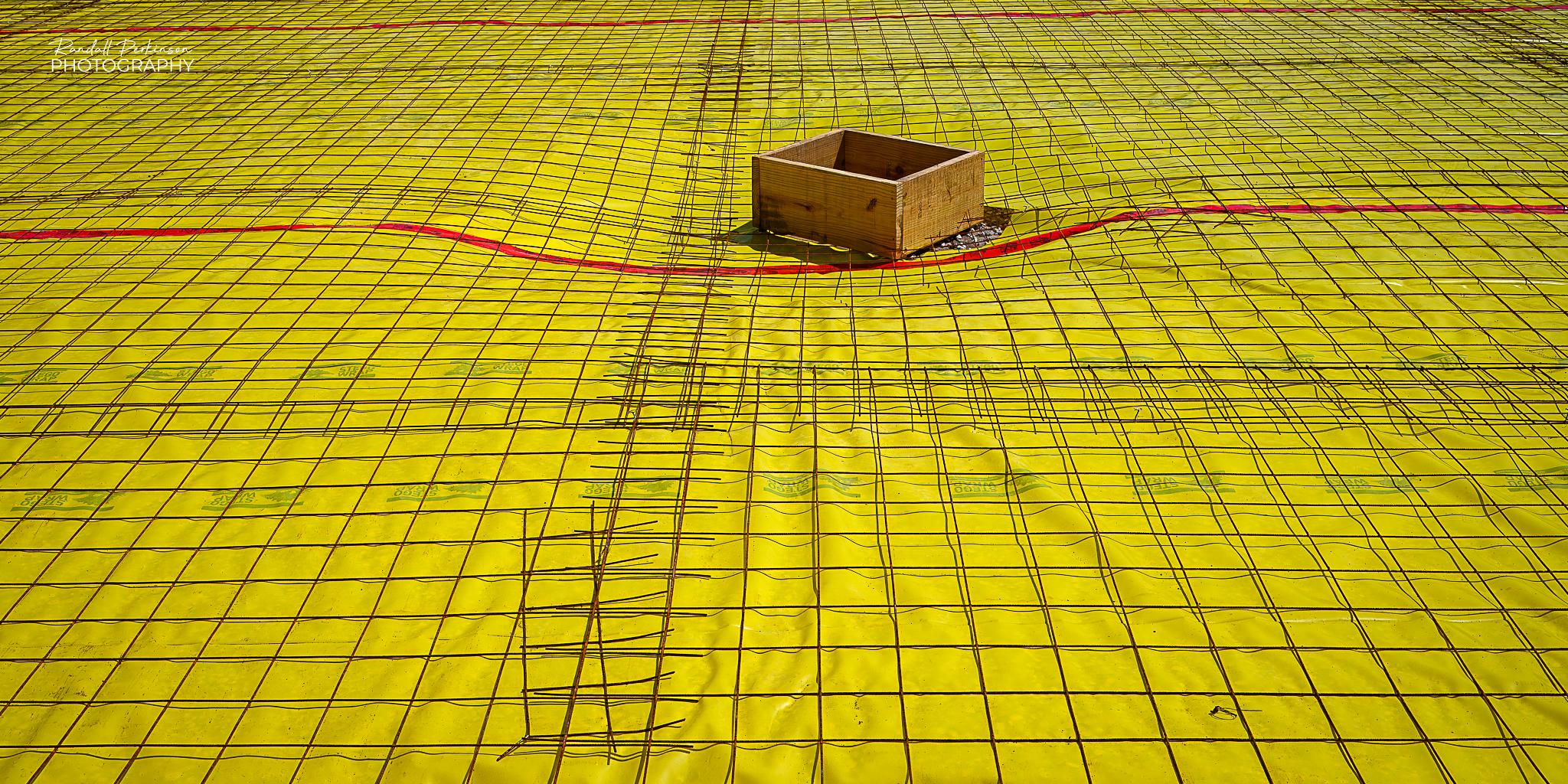 A view of an area being prepared for a concrete slab pour.  There is a plastic yellow vapor barrier covering the entire area with wire mesh reinforcement set on top of it.  In the middle is a wooden box for a block out in the slab.