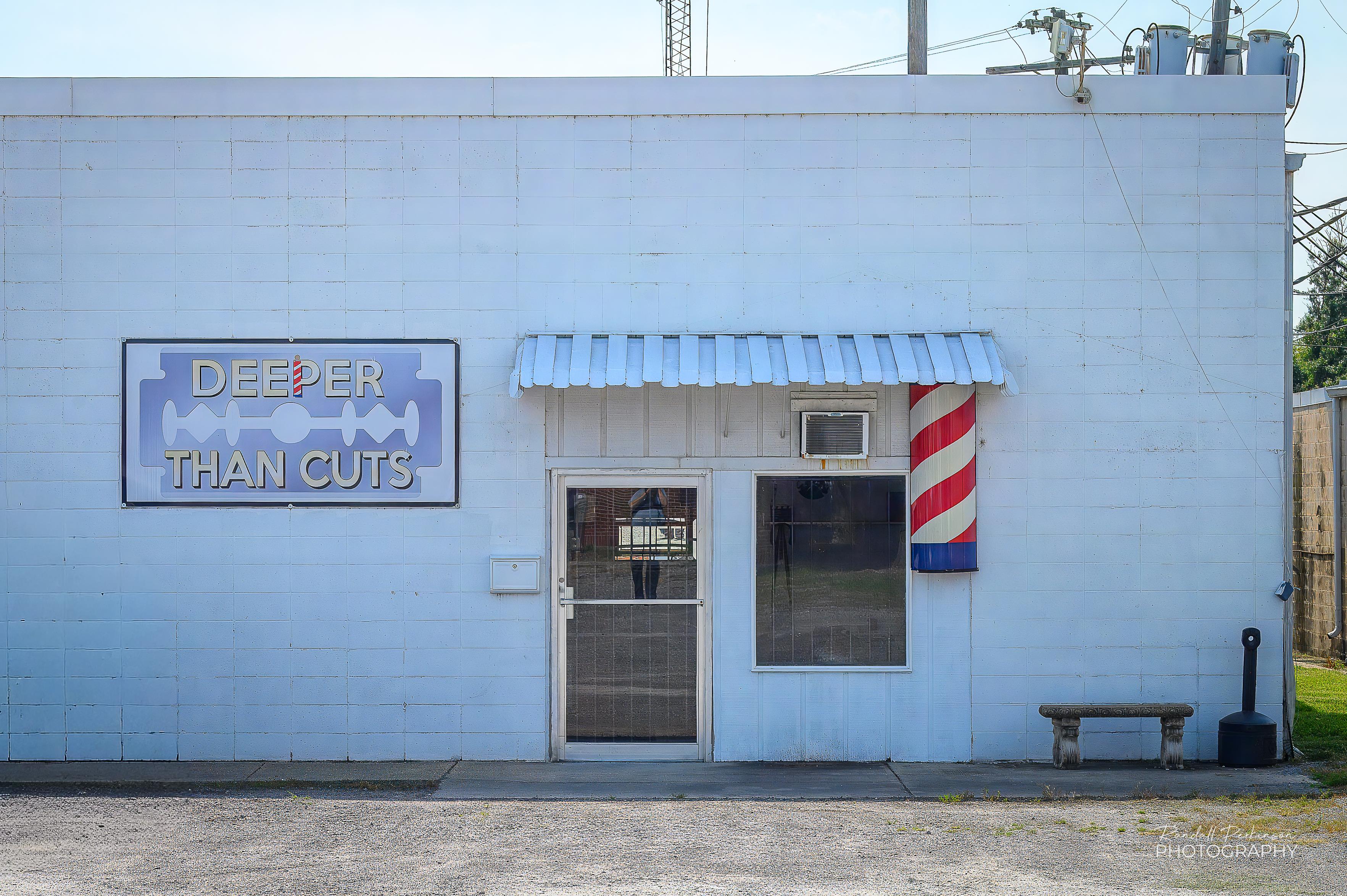 A barber shop located in a plain, white, one-story concrete block building.  There is a glass and metal door, one window, a partial barber pole and an awning over all three.  A sign shows a razor blade with with the words "DEEPER THAN CUTS" on it.
