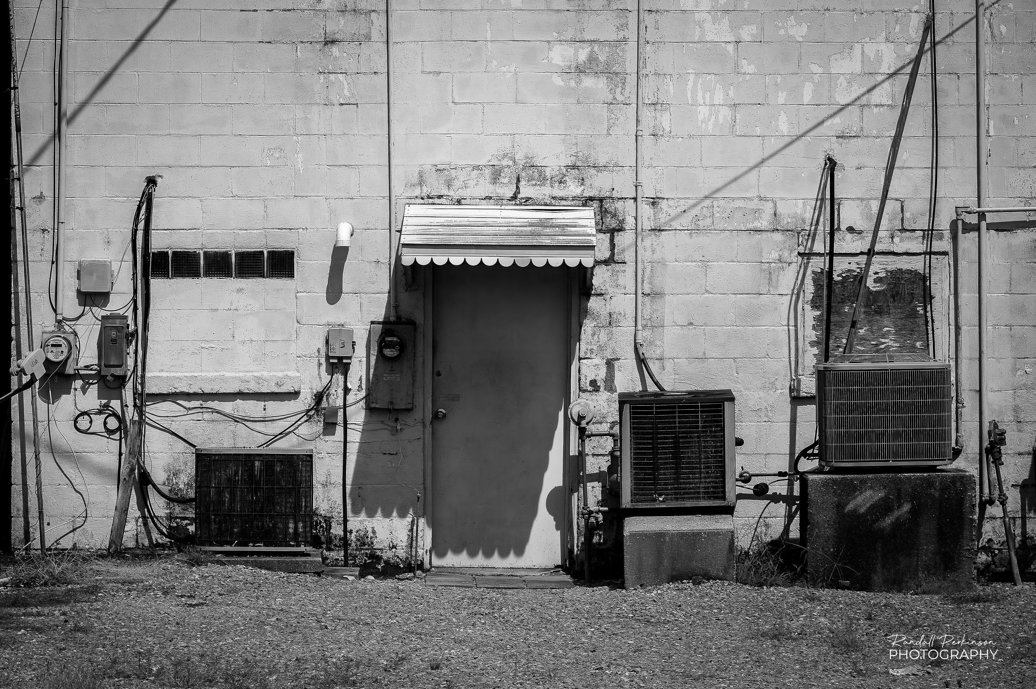 A view of the back of a concrete block building.  It has a single door with a small awning over it and three separate air conditioners sitting on either side of it.