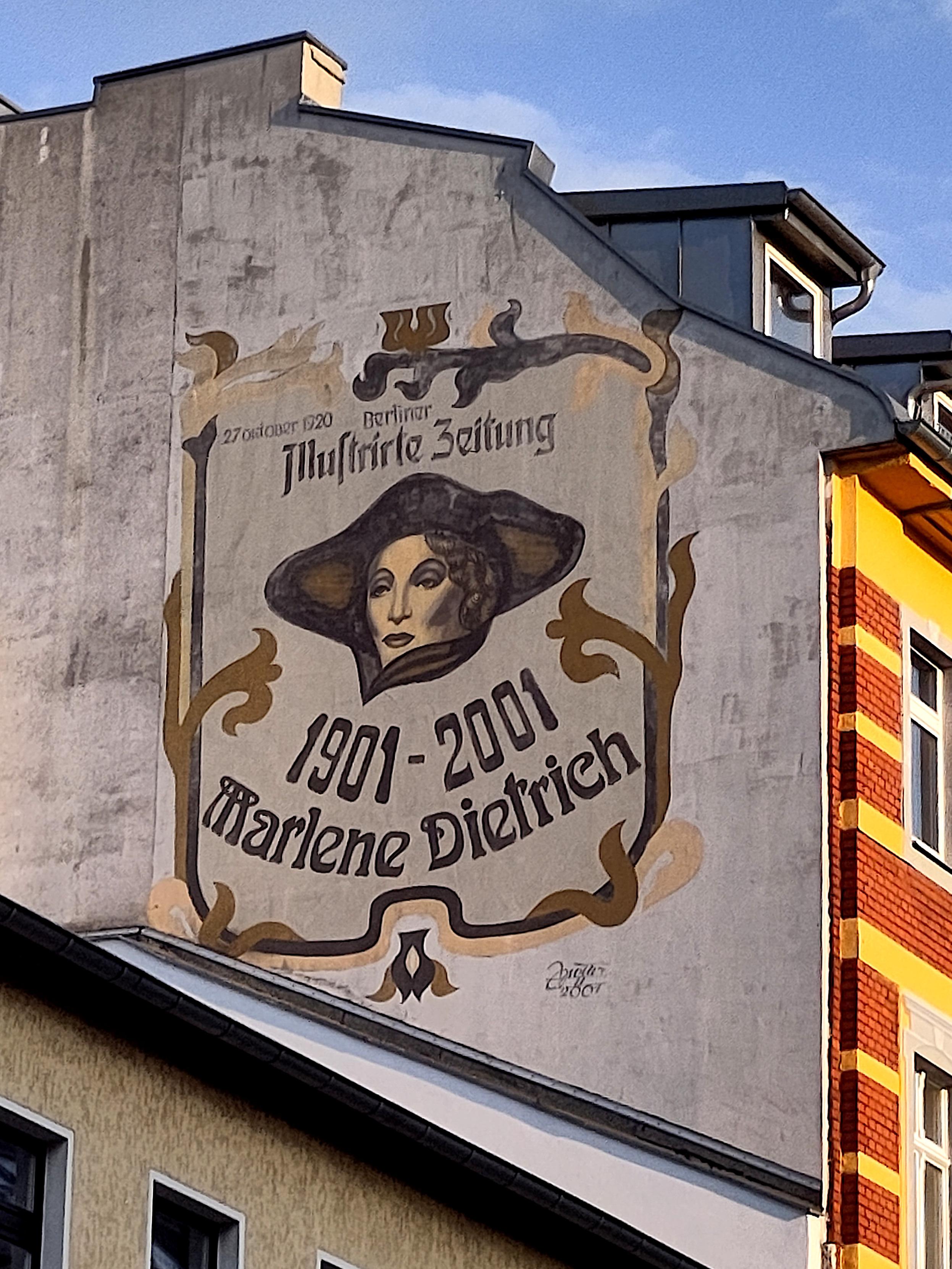A large mural painted on the grey firewall of a building, seen from below against a blue sky. In an Art Nouveau style with ornate golden-brown scrollwork framing, it shows the portrait of a woman's face beneath a wide-brimmed hat, rendered in muted grey and beige tones. Painted text reads "27 Oktober 1920 Berliner Illustrirte Zeitung" above the portrait and "1901–2001 Marlene Dietrich" below it in a decorative Gothic-style script, with a small signature and date in the lower corner. To the right stands a red-brick and yellow building; a lower rooftop is visible in the foreground.